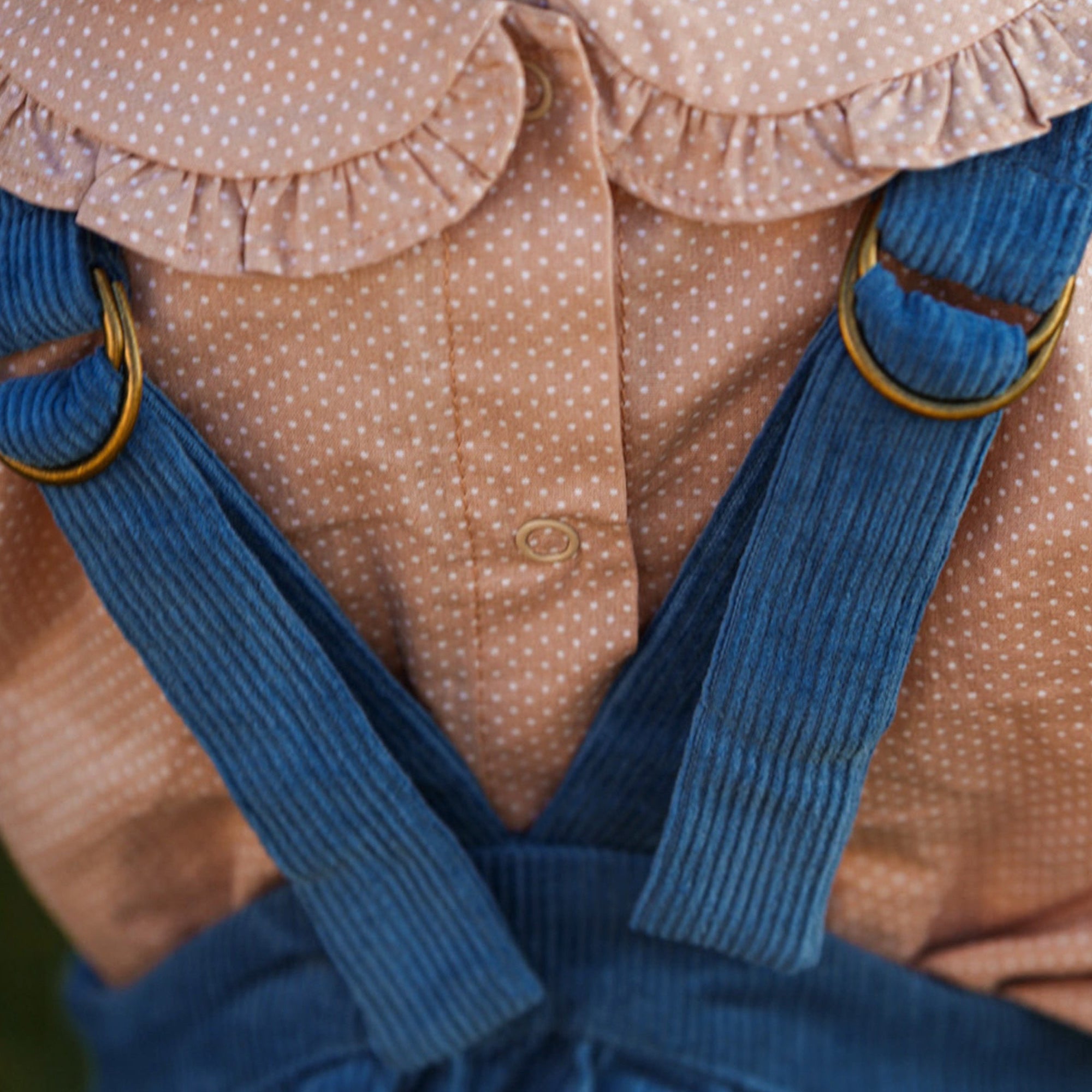Child wearing a beige blouse with ruffled collar and blue pinafore suspenders against a sunflower field.
