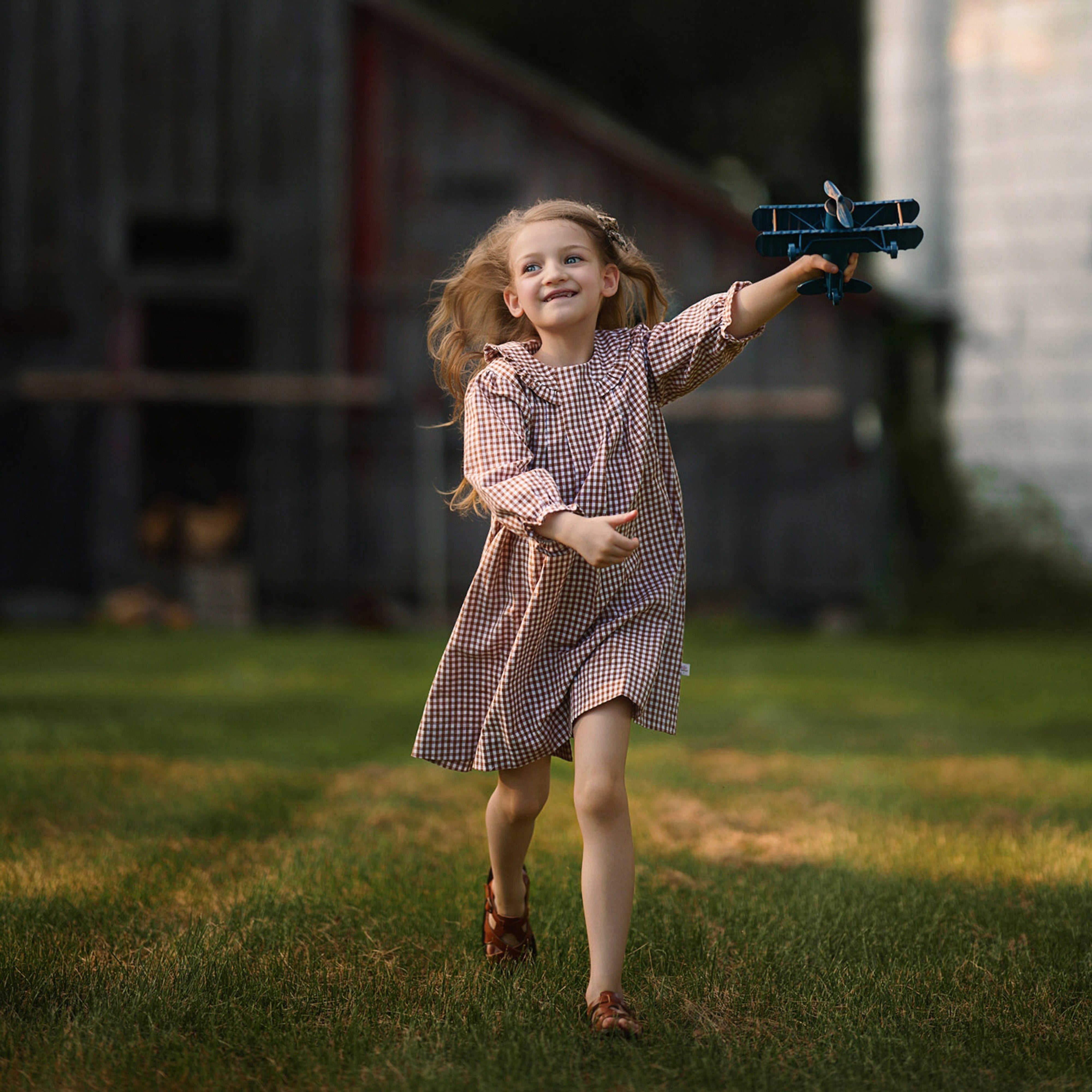 Young girl holding a toy airplane in a rural setting with a barn and trees in the background.