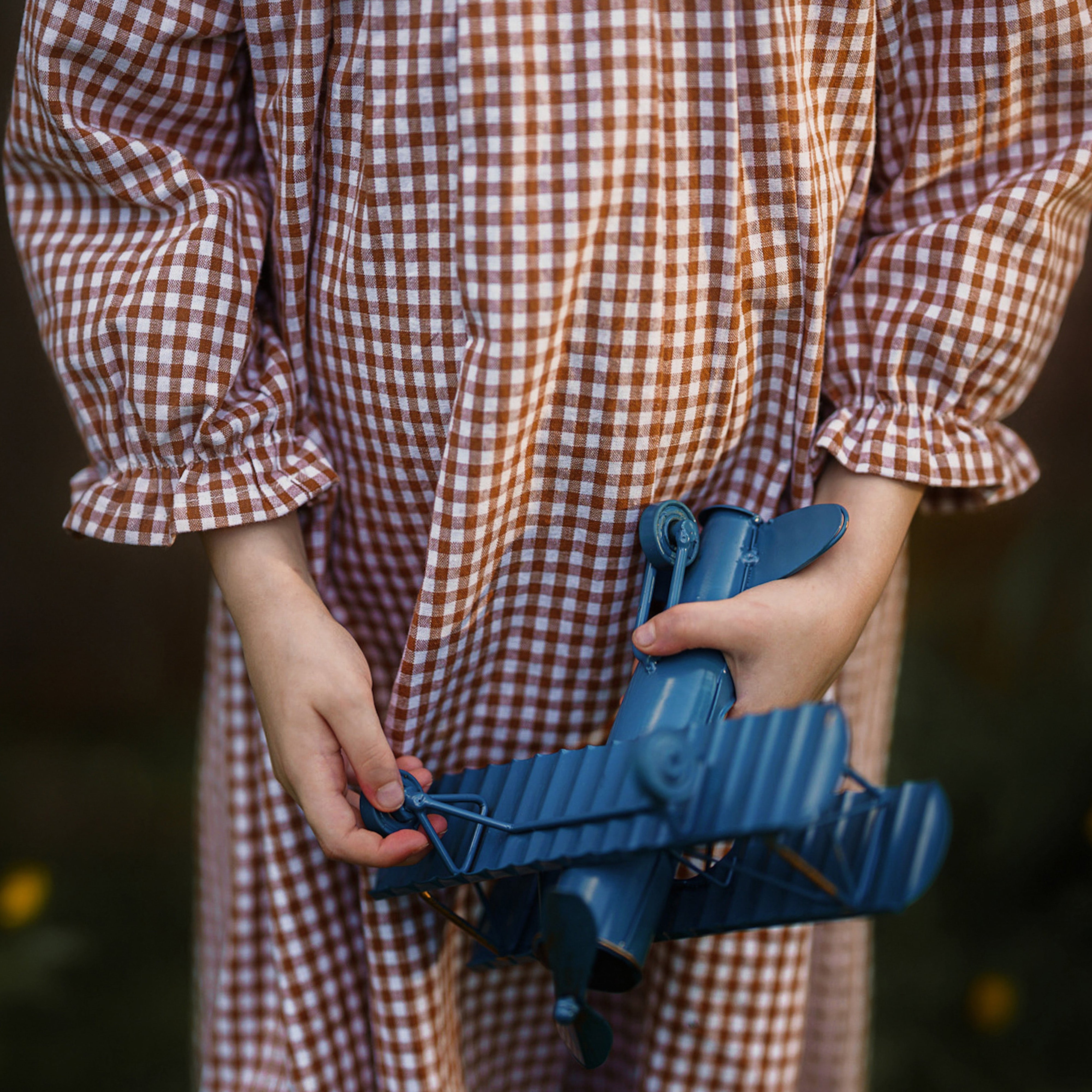 Girl wearing a checkered outfit holding a blue airplane toy with a blurred natural background
