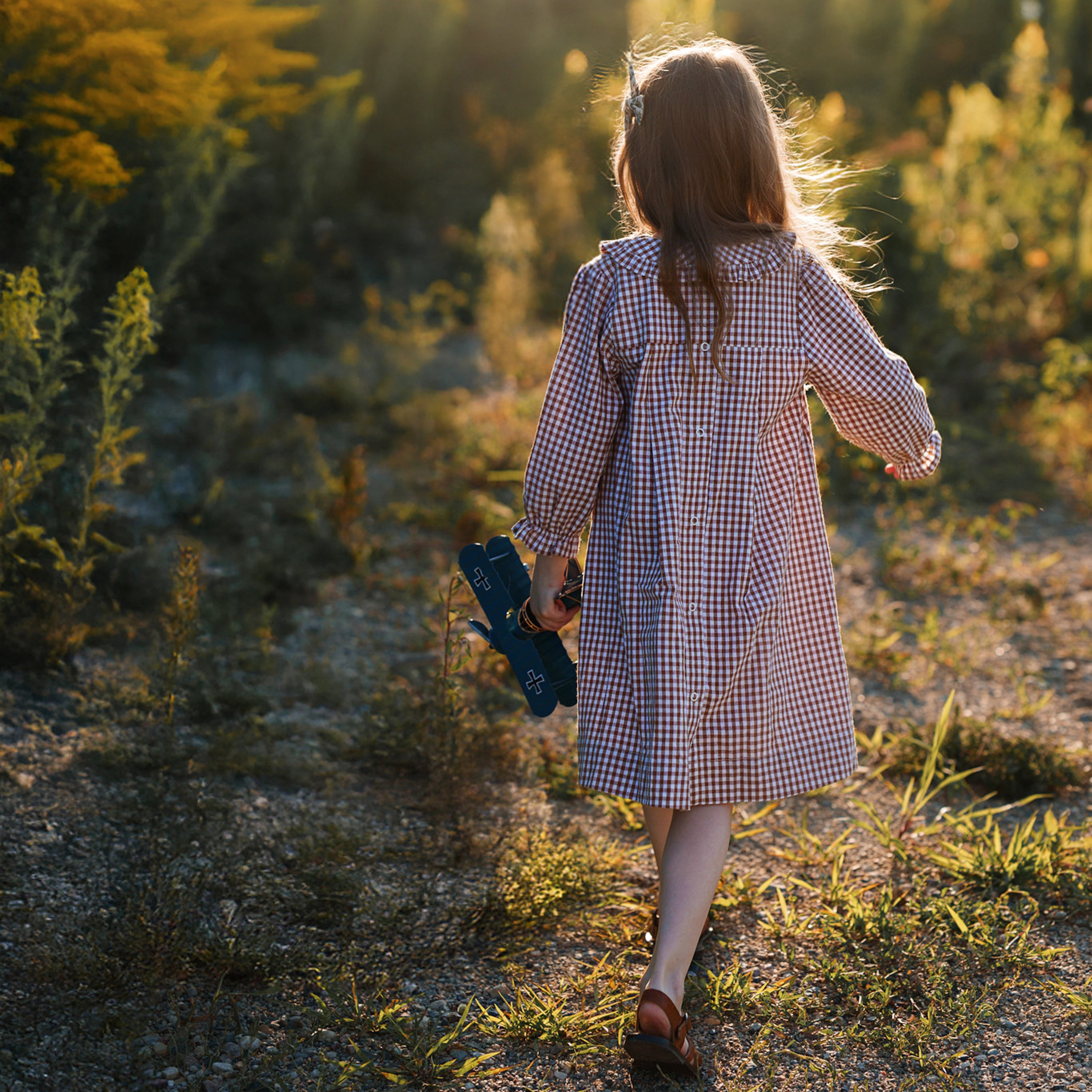 Girlwalking through a field with a checkered dress and airplane toy at sunset.