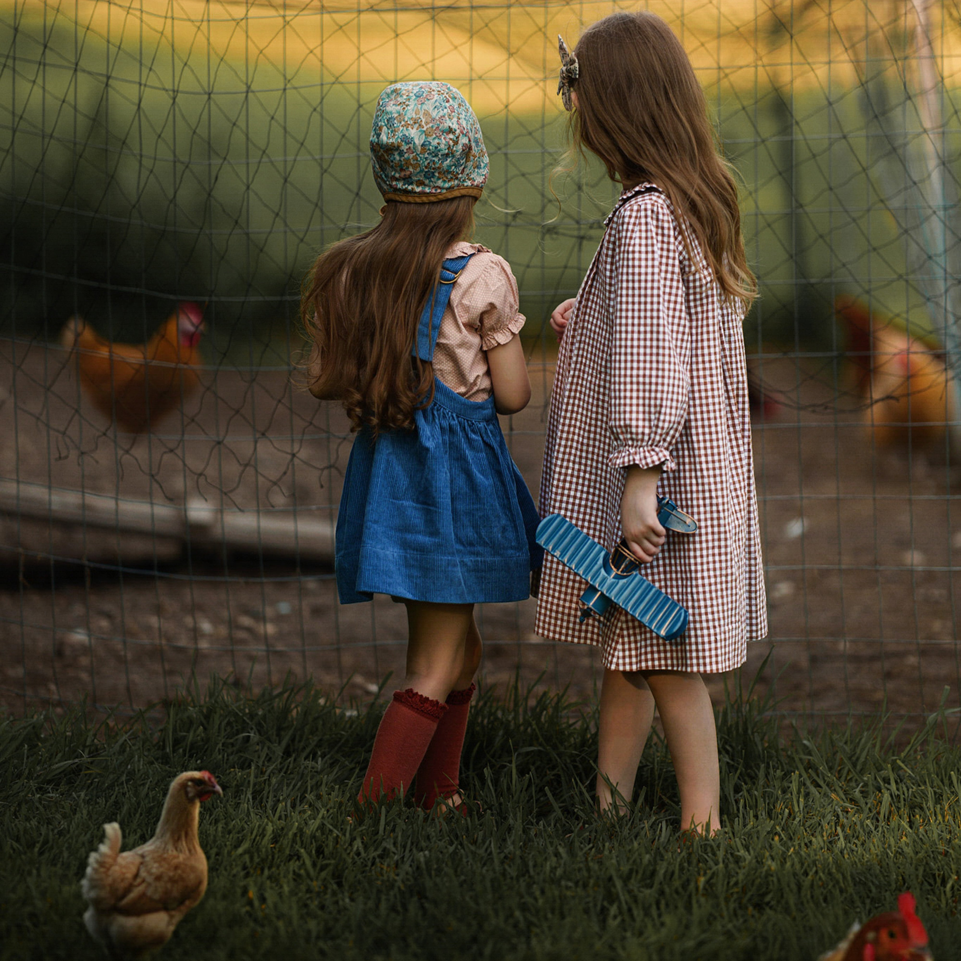 Two young girls standing in a grassy area with chickens around them, looking at a fence.