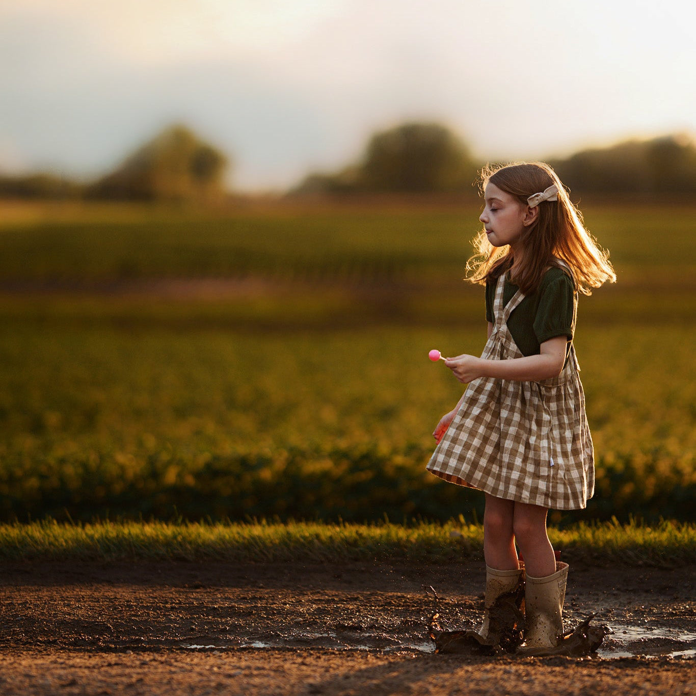 Young girl standing in a field with a sunset in the background in a checkered pinafore eating a lollipop
