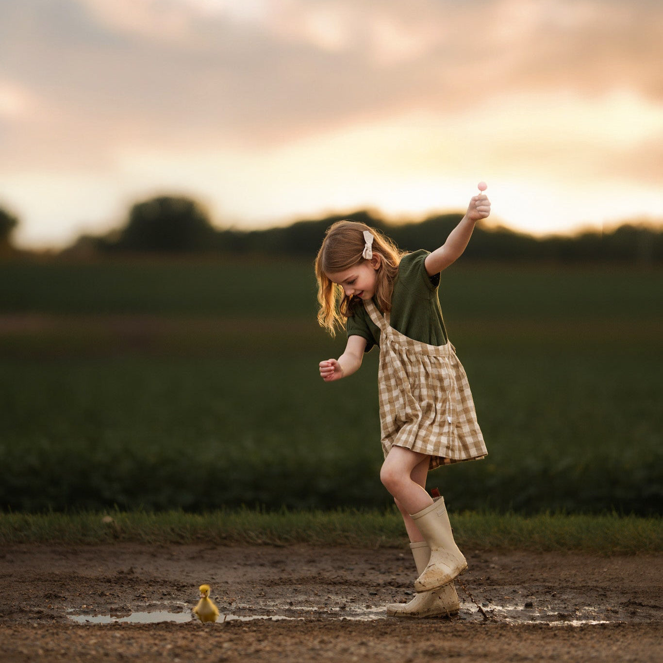 Child playing in a muddy puddle with a duckling at sunset.