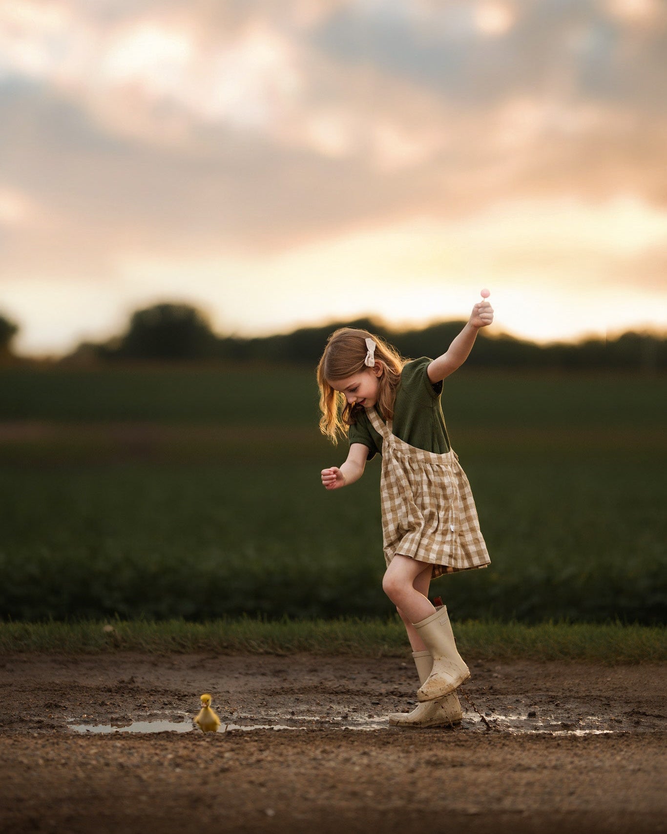 Child playing in a muddy puddle with a duckling at sunset.