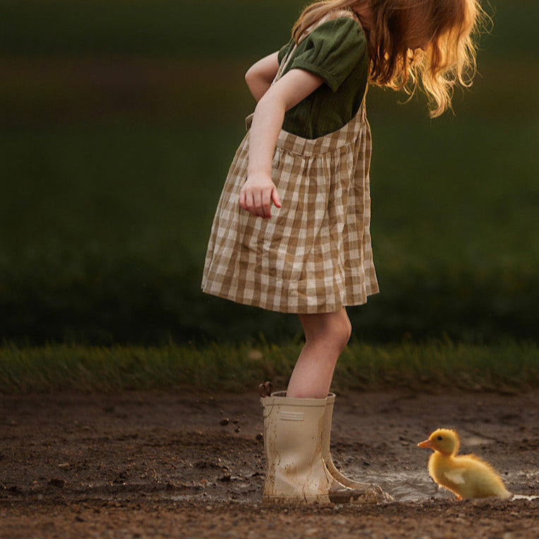 Young girl in checkered dress and boots standing next to a duckling in a field at sunset.