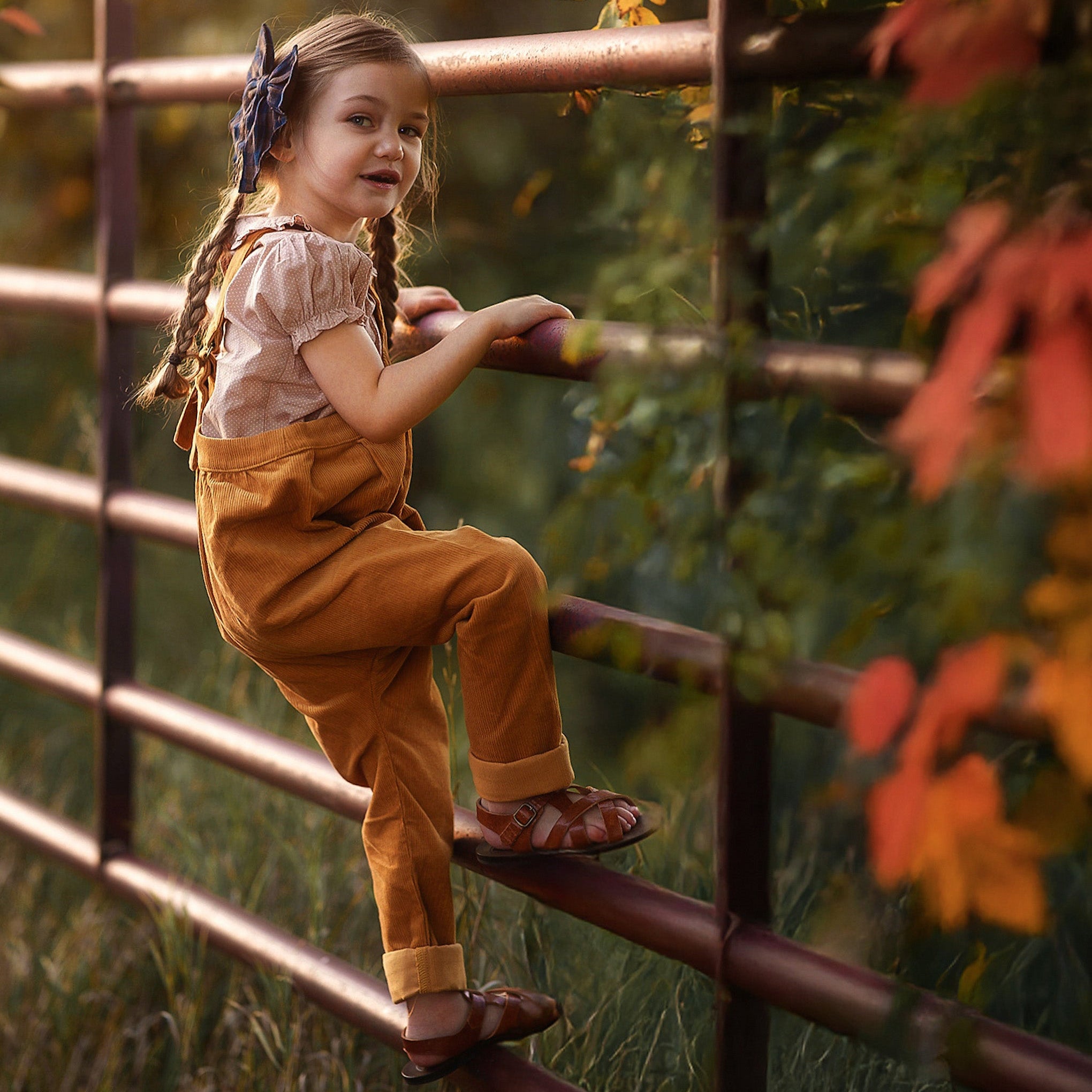 Young girl in orange overalls climbing a metal fence with autumn leaves in the background