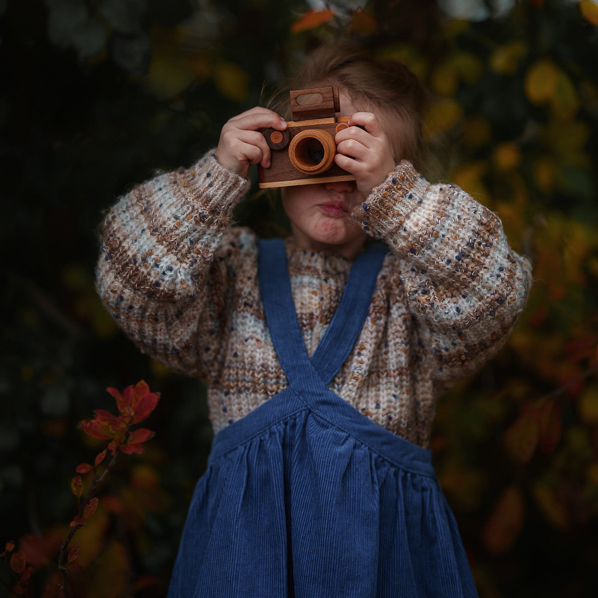 Child holding a vintage camera in a forest setting wearing a blue corduroy pinafore.