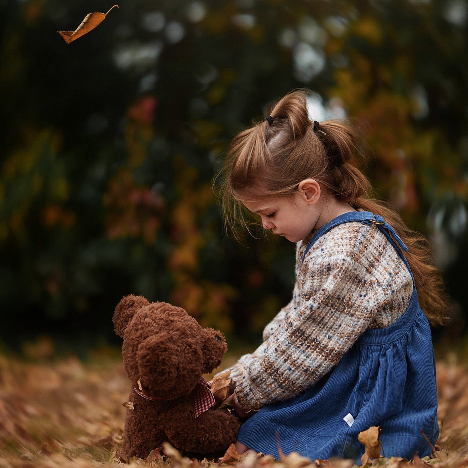 Child holding a teddy bear in an outdoor setting with trees and leaves in a blue corduroy pinafore.