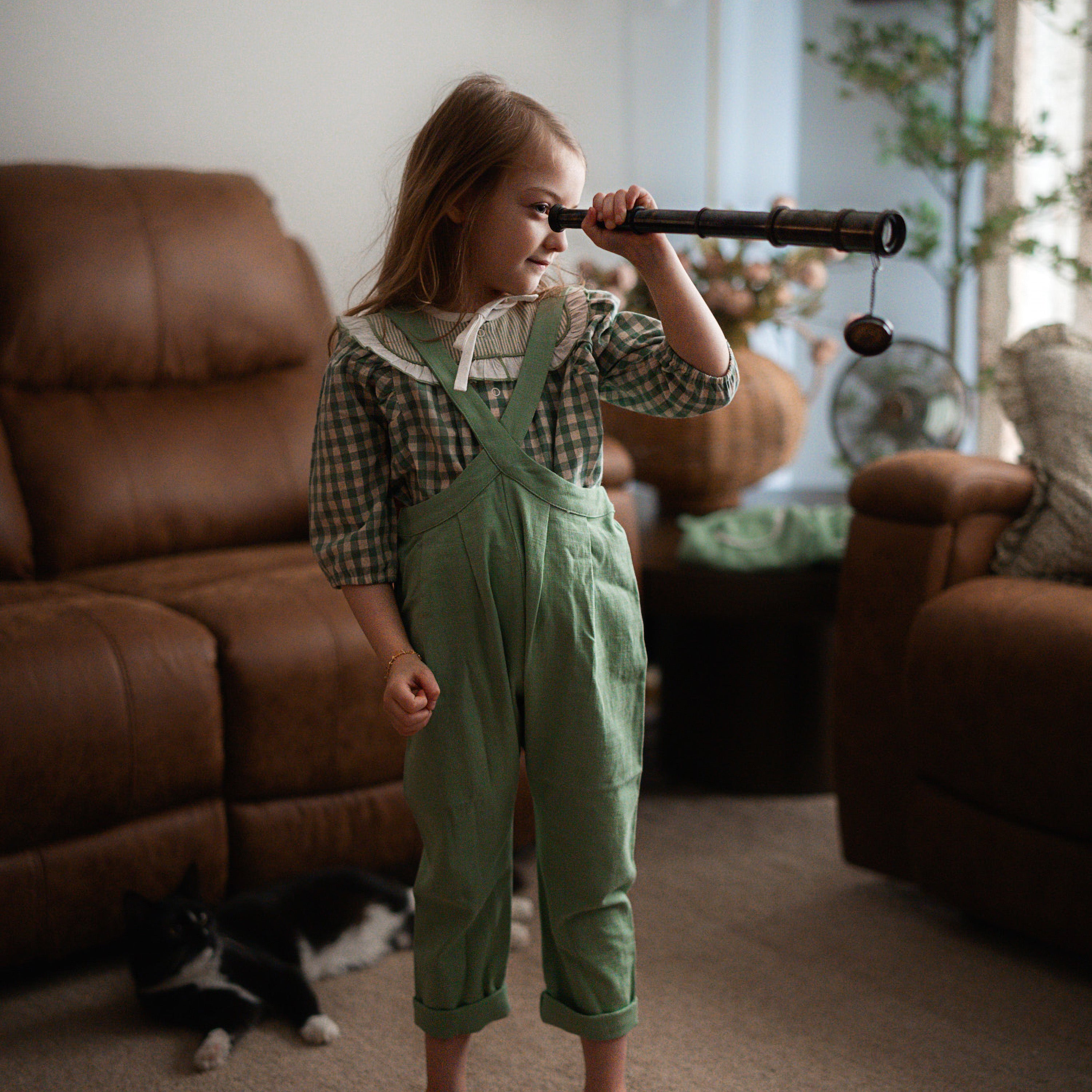 Child in green overalls holding a toy telescope in a living room.