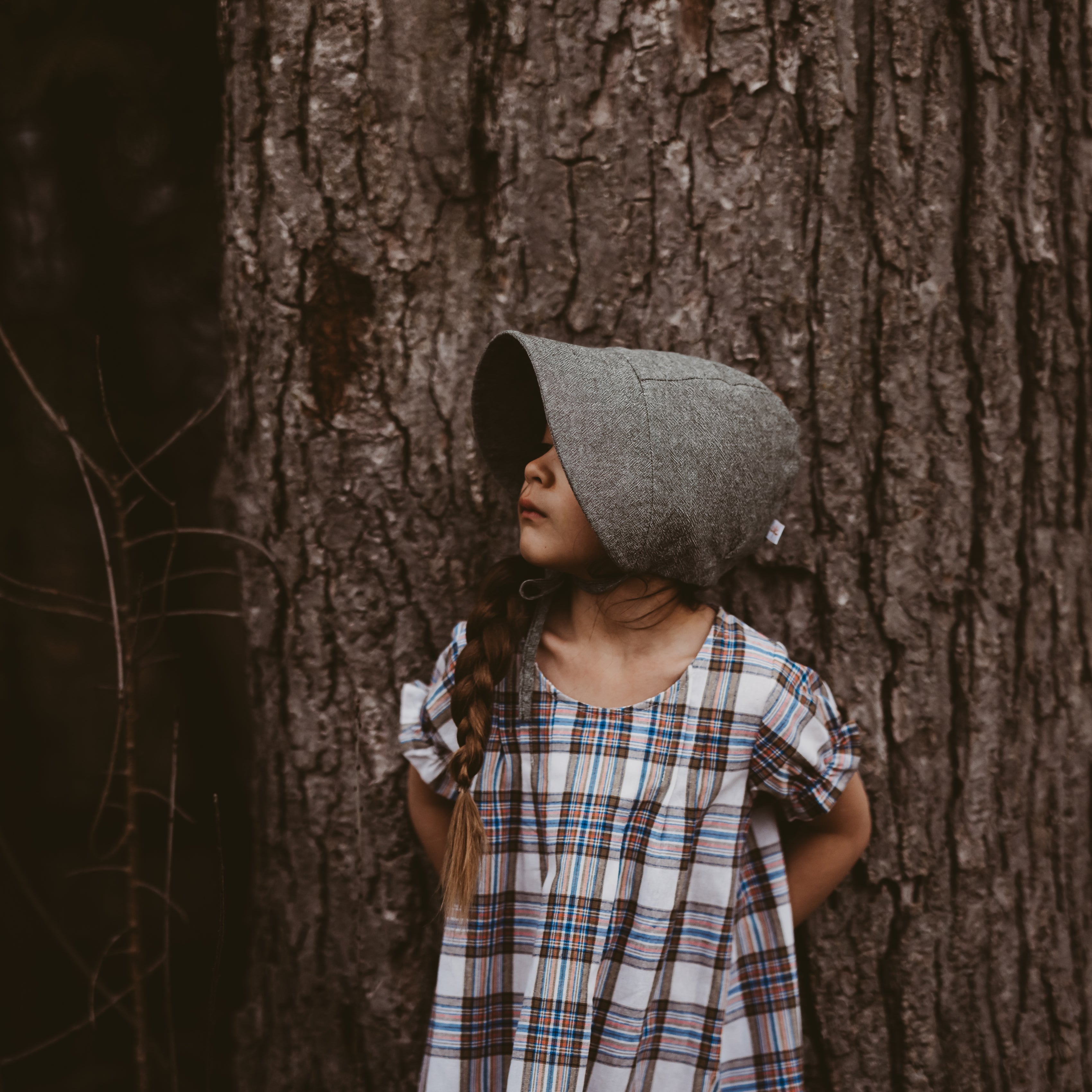 Girl in a plaid dress and green bonnet leaning against a tree