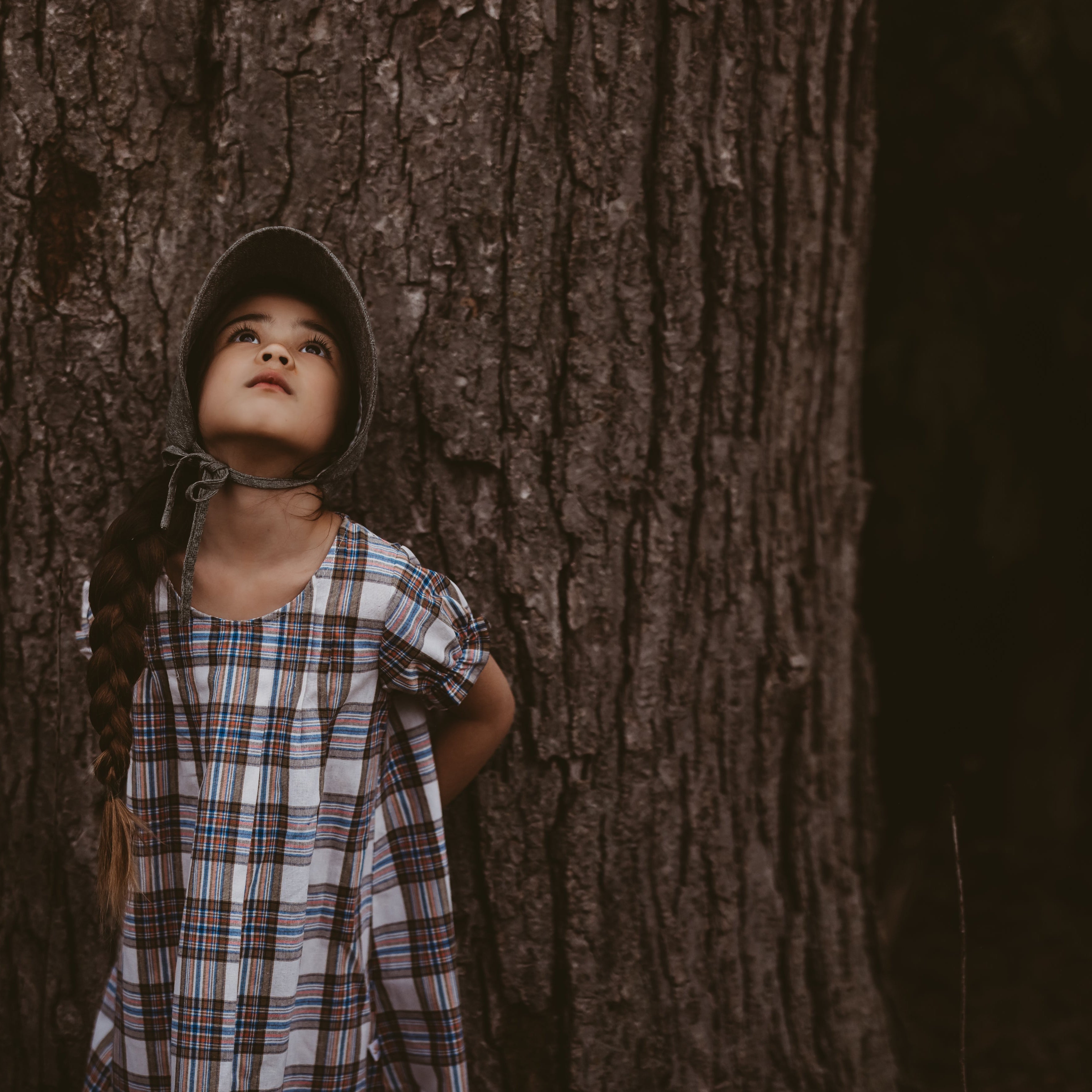 Girl in a plaid dress and green bonnet looking up leaning against a tree