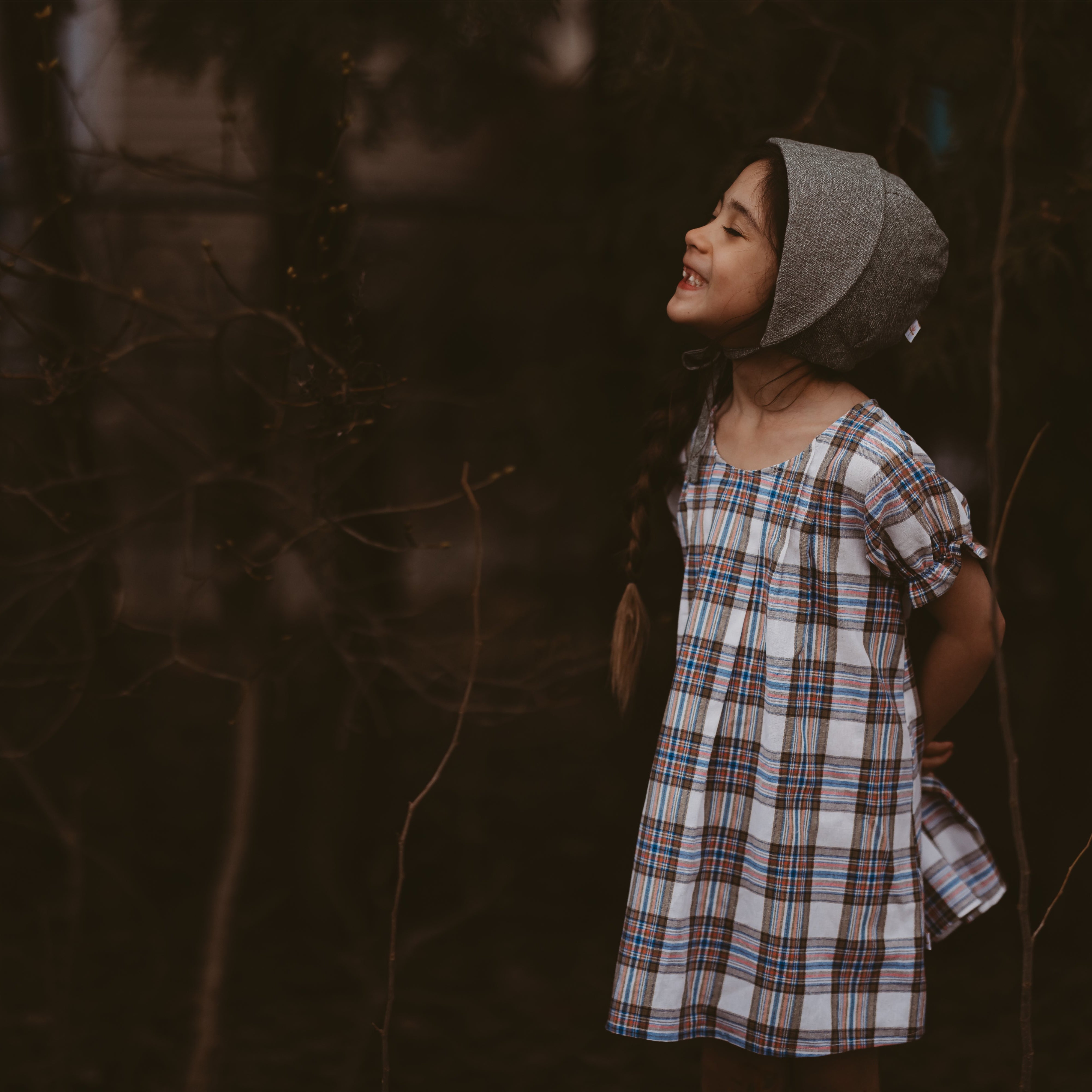 Girl wearing a plaid dress and a bonnet standing in a dark, forest-like setting.
