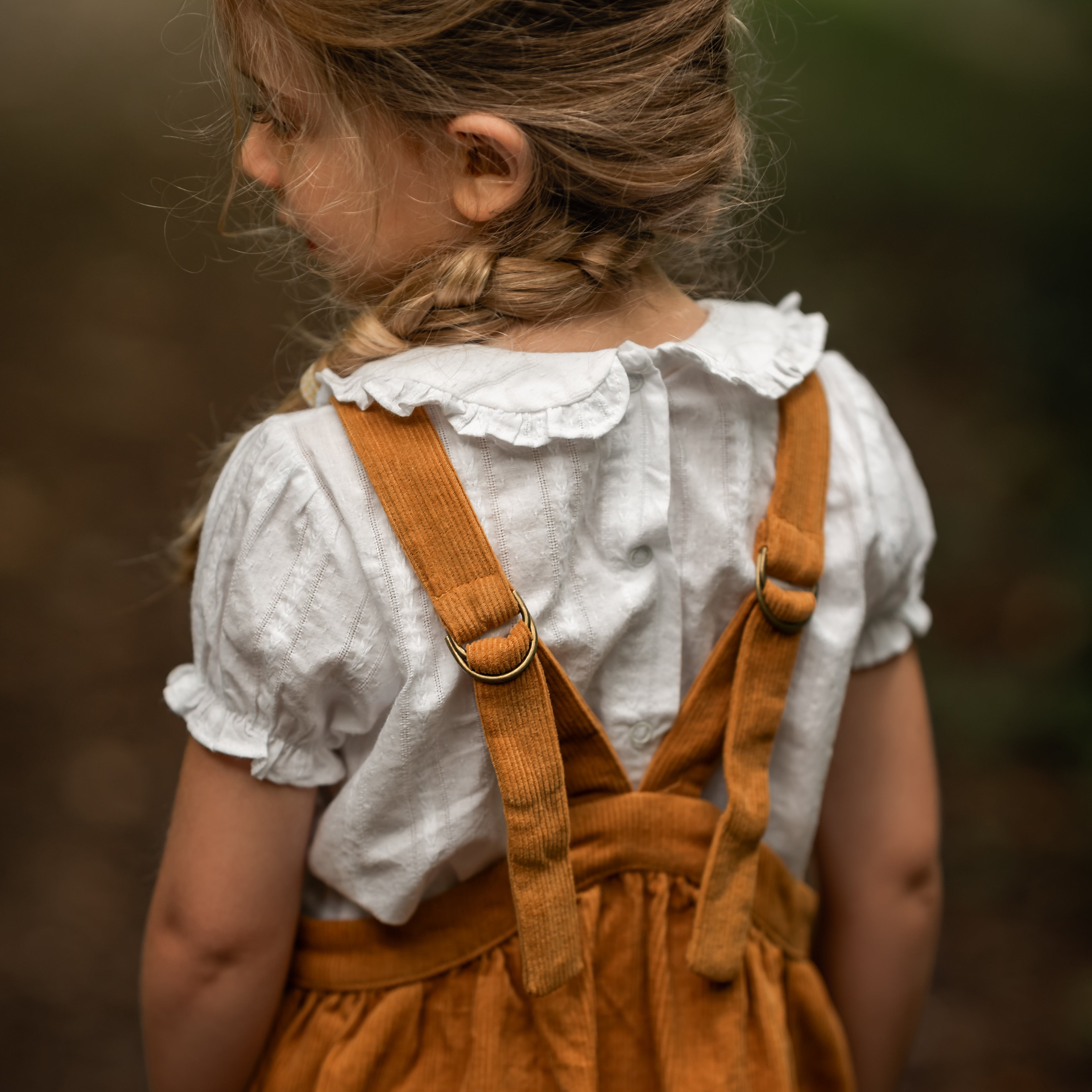 Child wearing a white blouse with a ruffled collar and mustard pinafore dress against a blurred natural background
