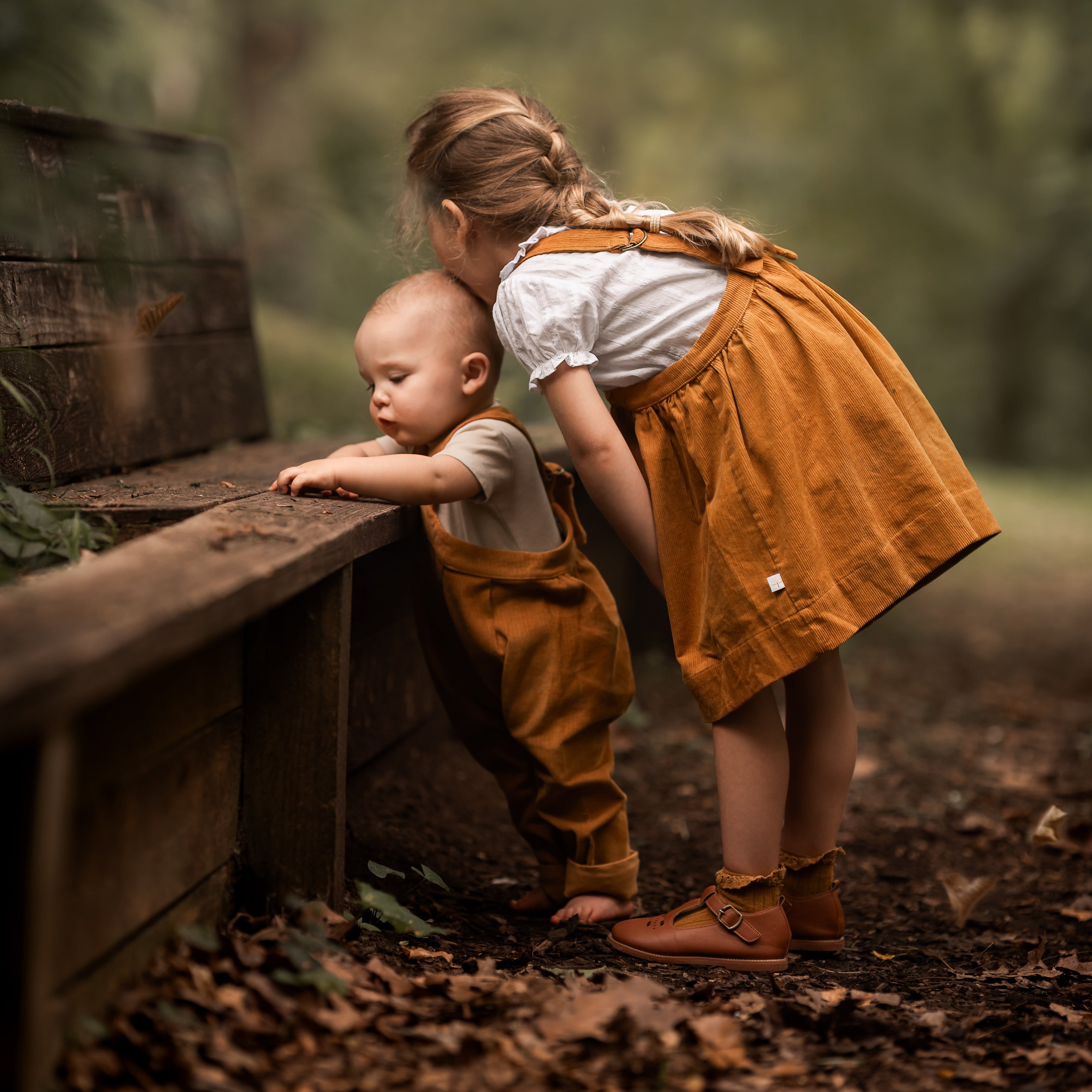 Two children in matching mustard corduroy outfits playing in a forest with leaves falling.