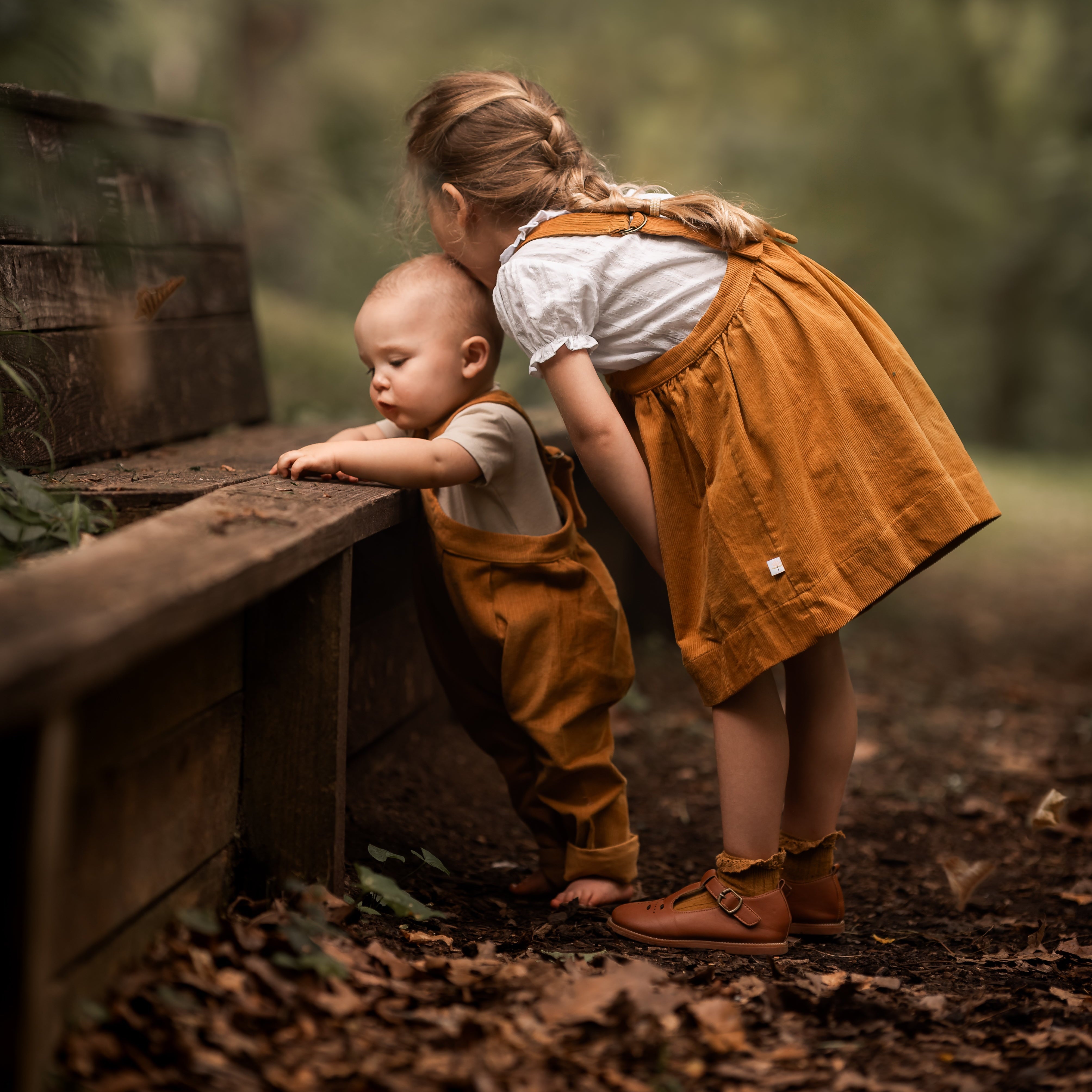 Two children in matching mustard corduroy outfits playing in a forest with leaves falling.