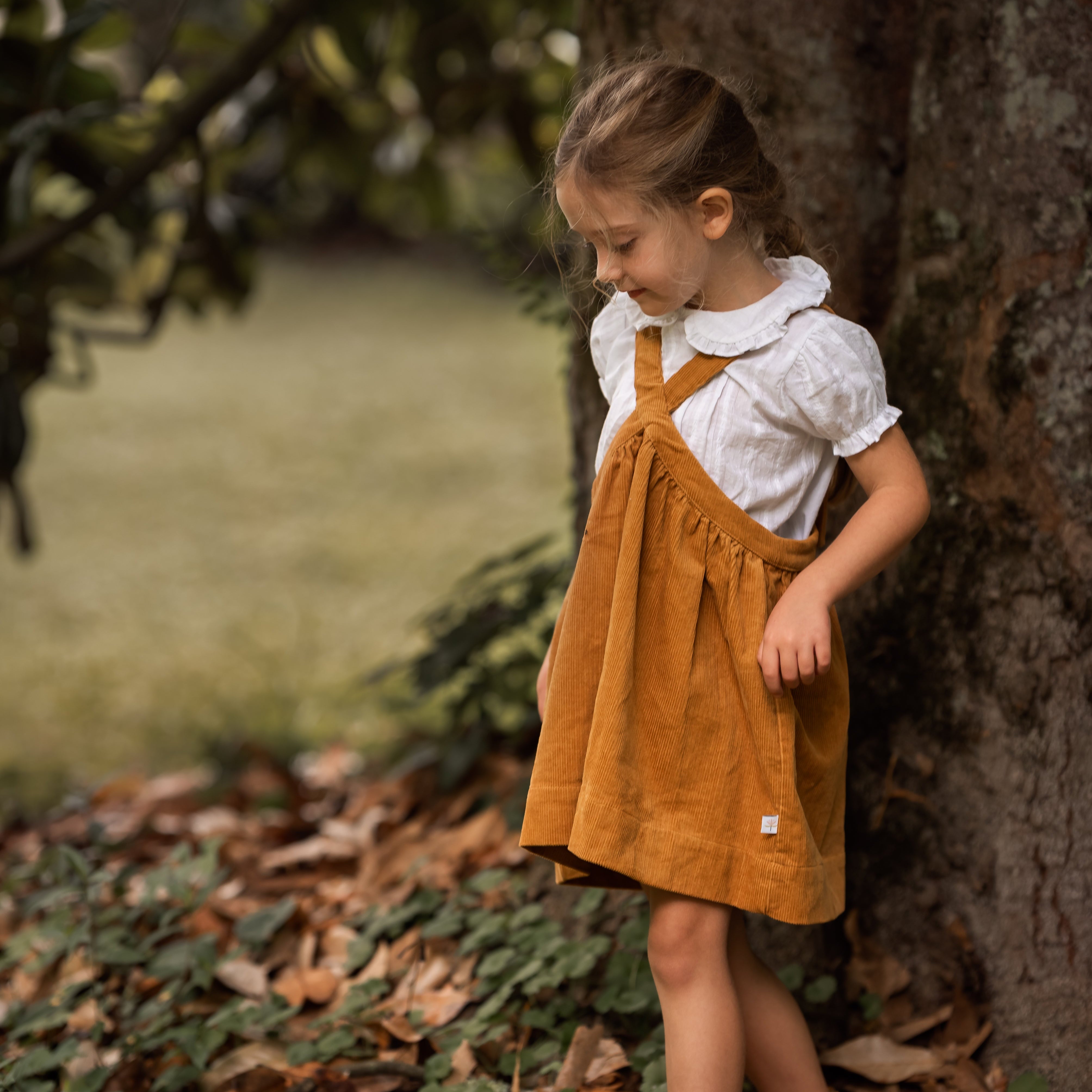 Young girl in a mustard pinafore dress standing next to a tree in a natural setting.