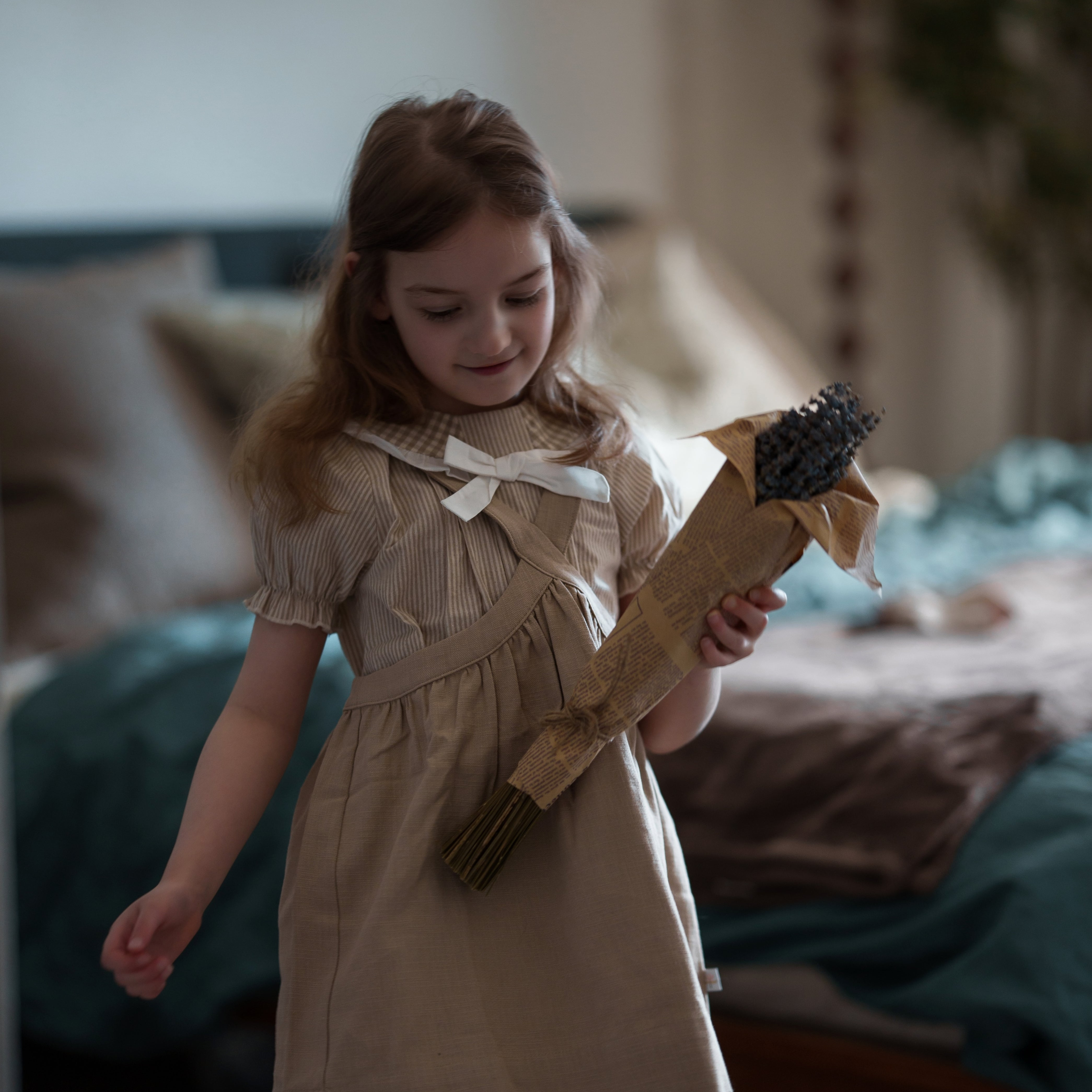 Young girl in a beige dress holding a toy broom in a bedroom setting