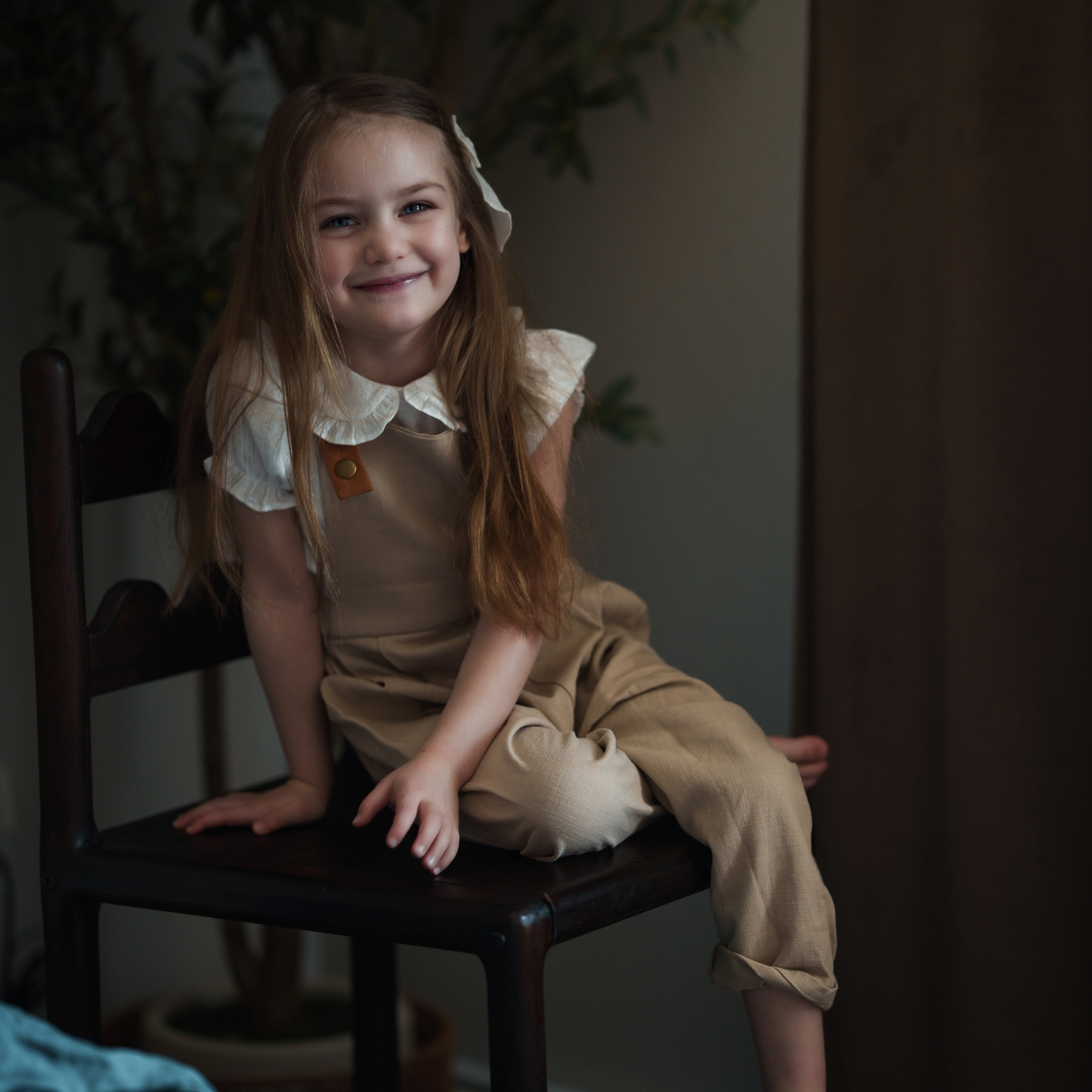 Young girl in a beige outfit sitting on a chair in a dimly lit room.