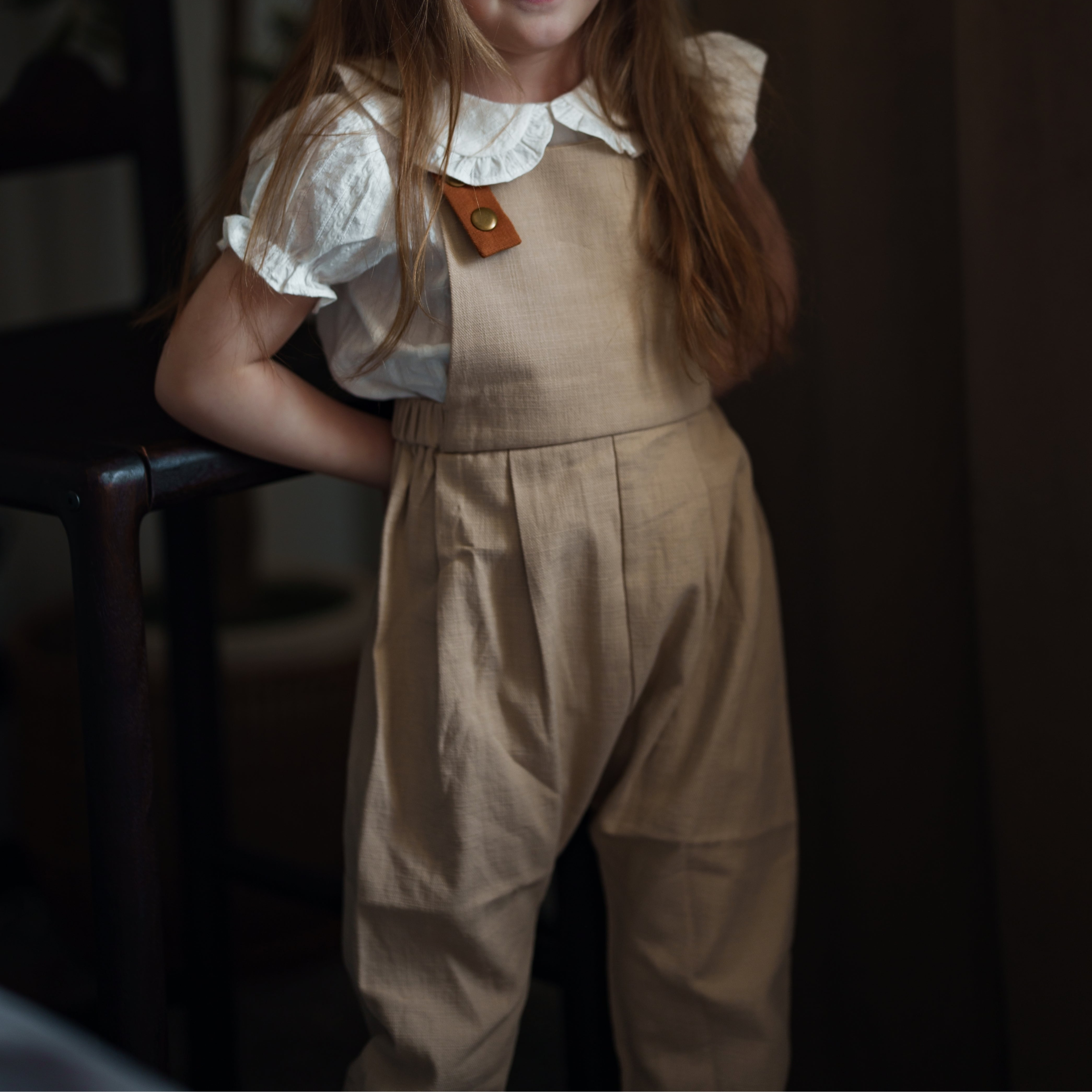 Young girl wearing a beige dress with a white collar and brown hair, standing indoors.