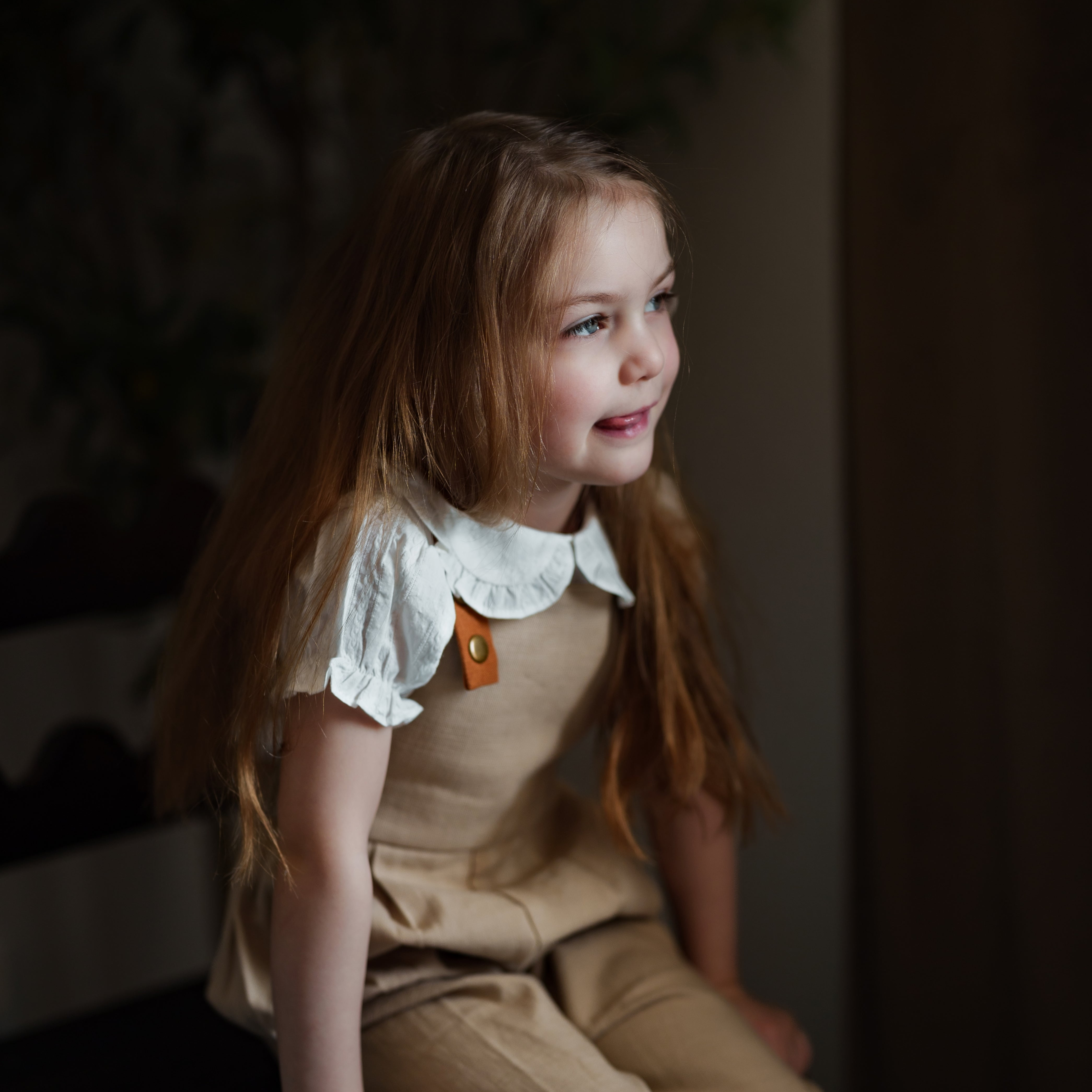 Young girl in a beige outfit with a white lace collar sitting on a dark surface.