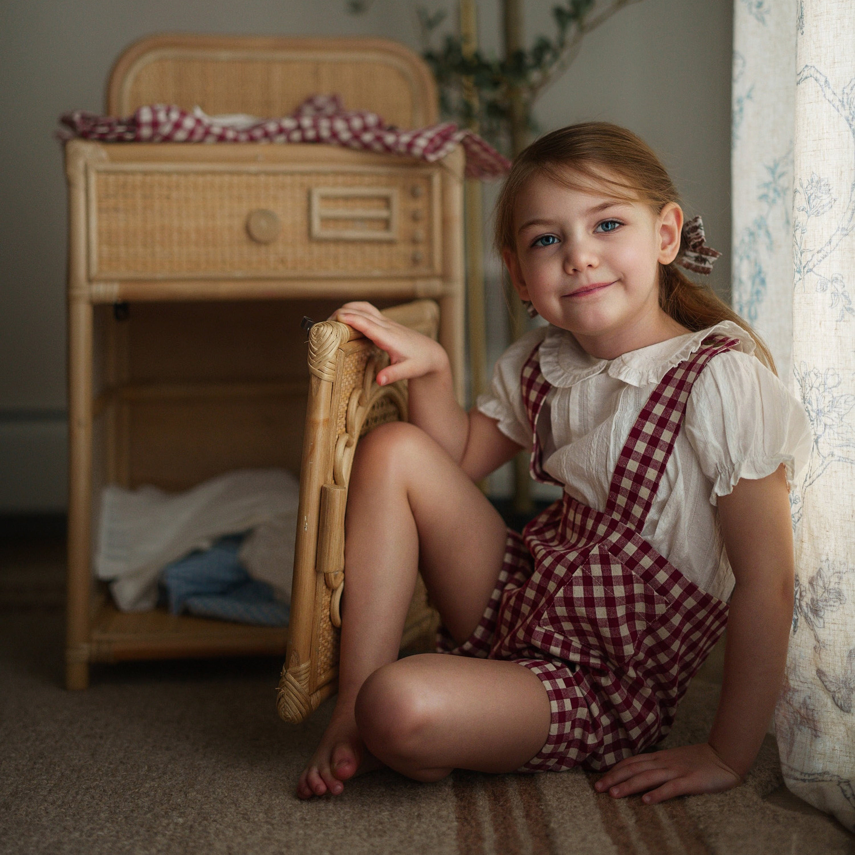 Young girl in a checkered dress sitting on a chair in a home setting.