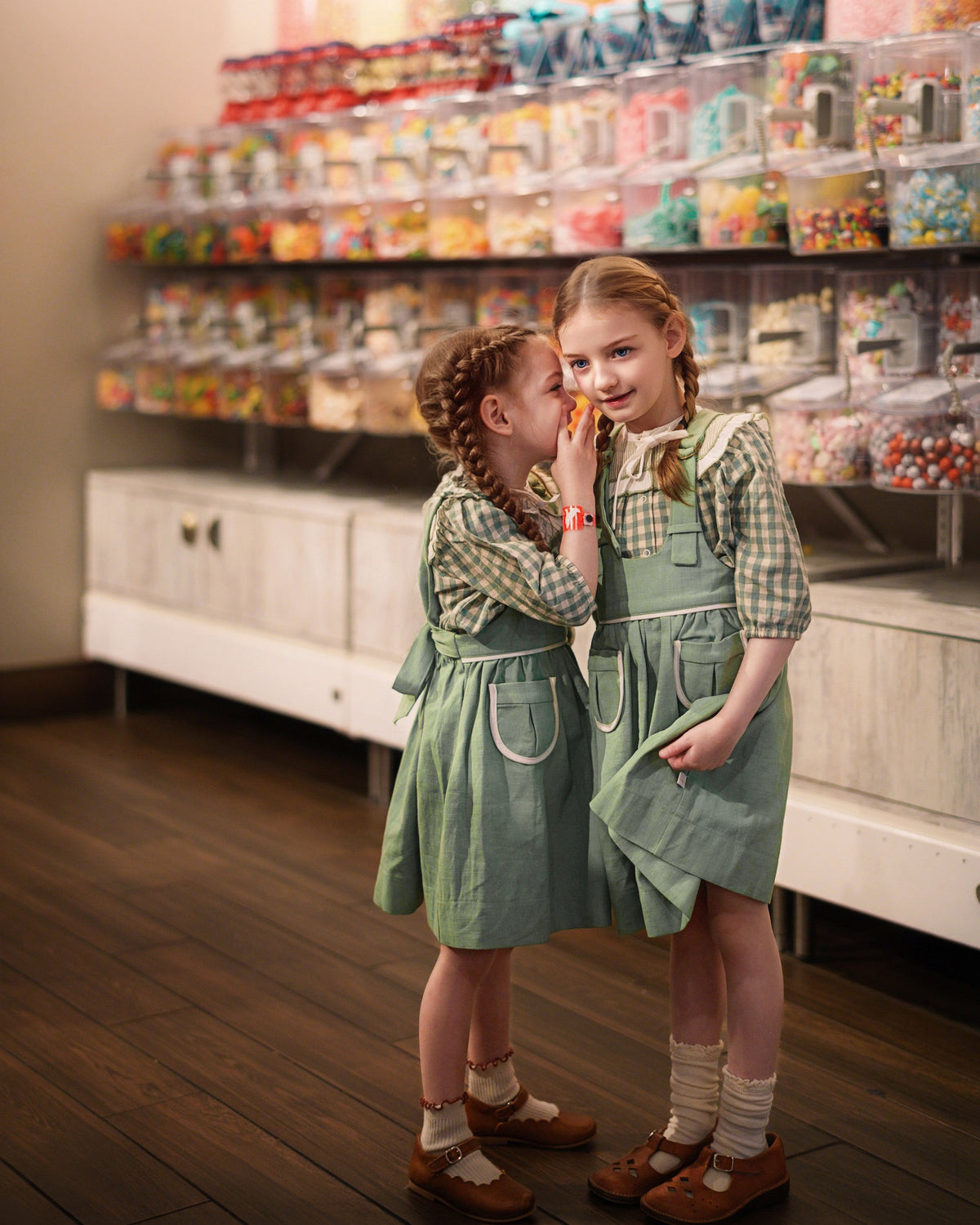 Two young girls in matching green pinafore dresses and plaid shirts pose together in front of a large candy display filled with colorful gummy candies and jars.