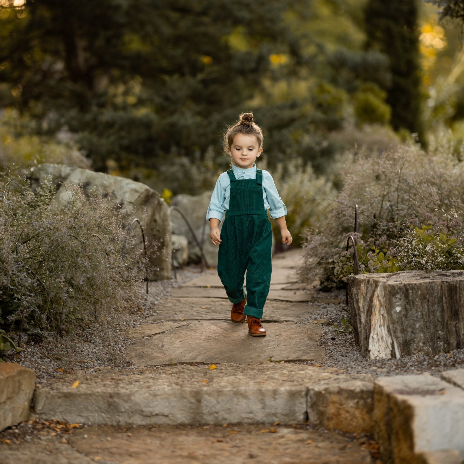 A 3-year-old model boy walking outdoors in a classic light blue Henley shirt with white Swiss dots, crafted from natural, sustainable, and high-quality materials. He is paired with spruce corduroy long overalls that enhance the outfit's texture and warmth. This relaxed pose highlights the stylish yet comfortable nature of the ensemble, perfect for everyday adventures.