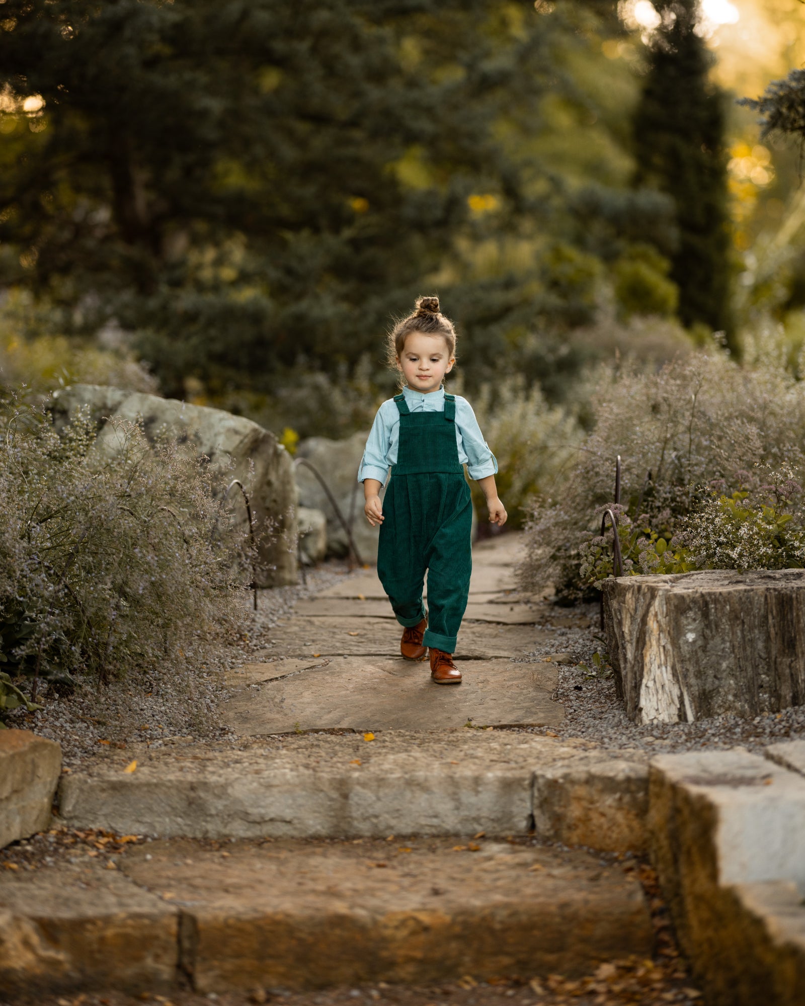 A 3-year-old model boy walking outdoors in a classic light blue Henley shirt with white Swiss dots, crafted from natural, sustainable, and high-quality materials. He is paired with spruce corduroy long overalls that enhance the outfit's texture and warmth. This relaxed pose highlights the stylish yet comfortable nature of the ensemble, perfect for everyday adventures.