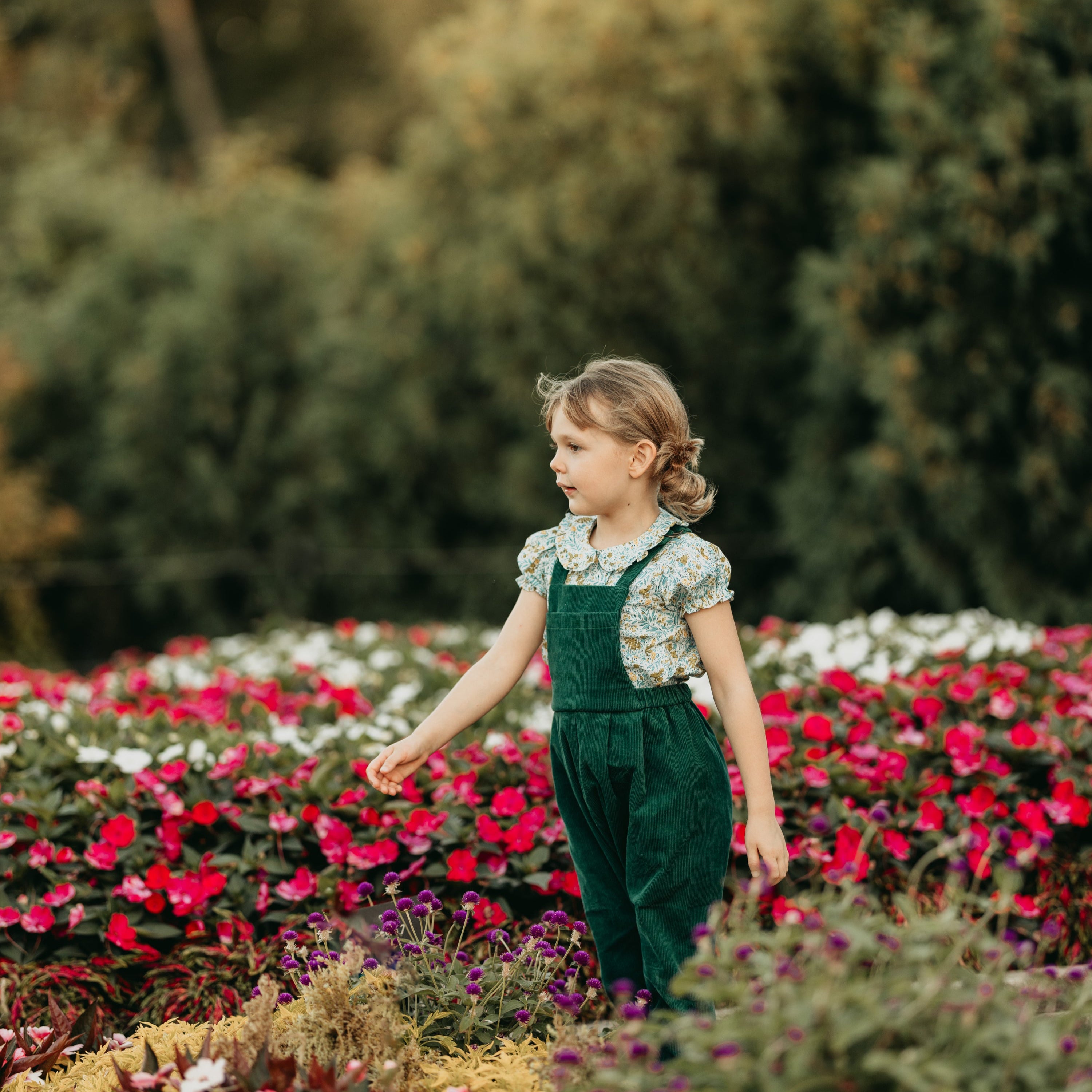 Young girl in green overalls standing among colorful flowers in a garden.