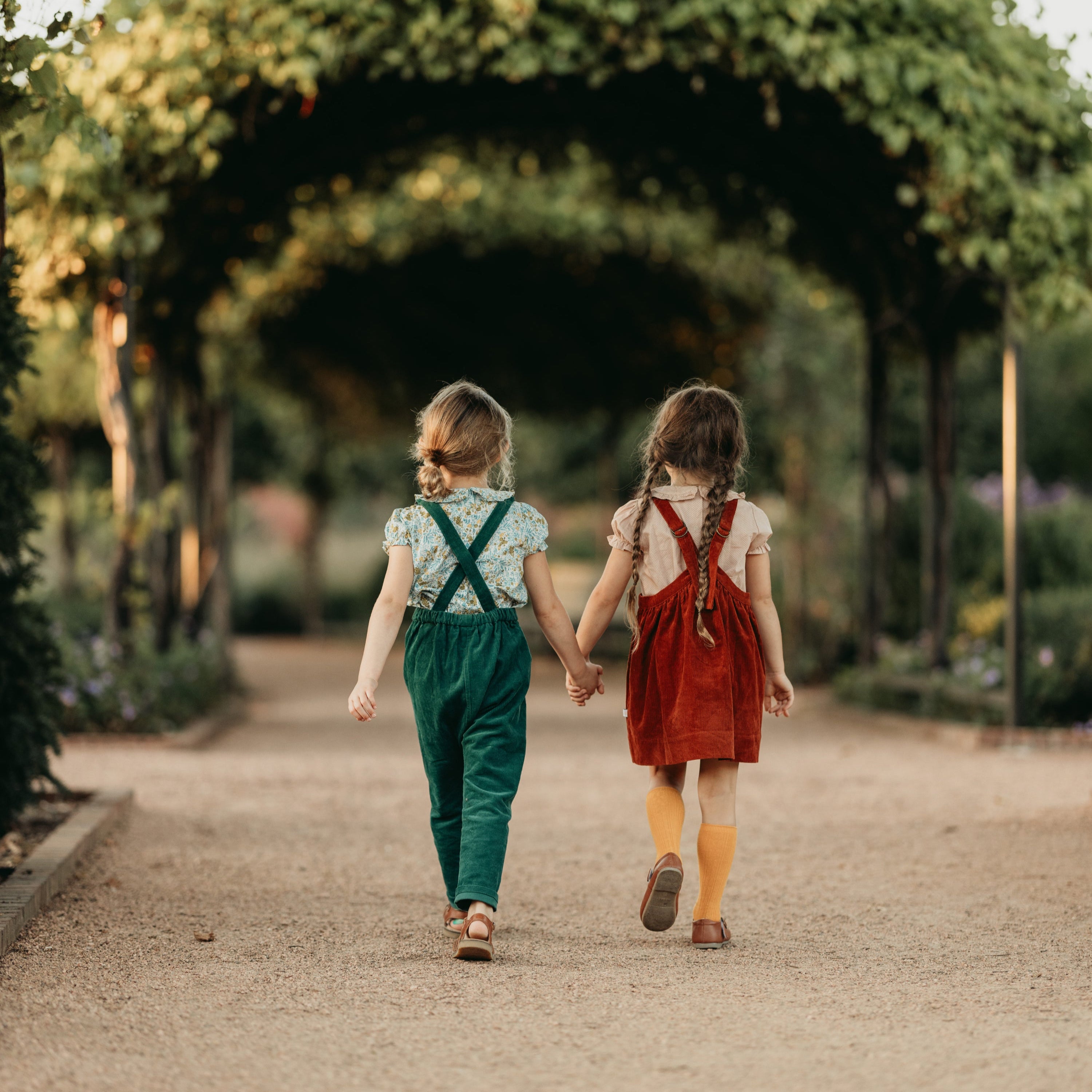 Two girls holding hands and walking away on a path with greenery around.