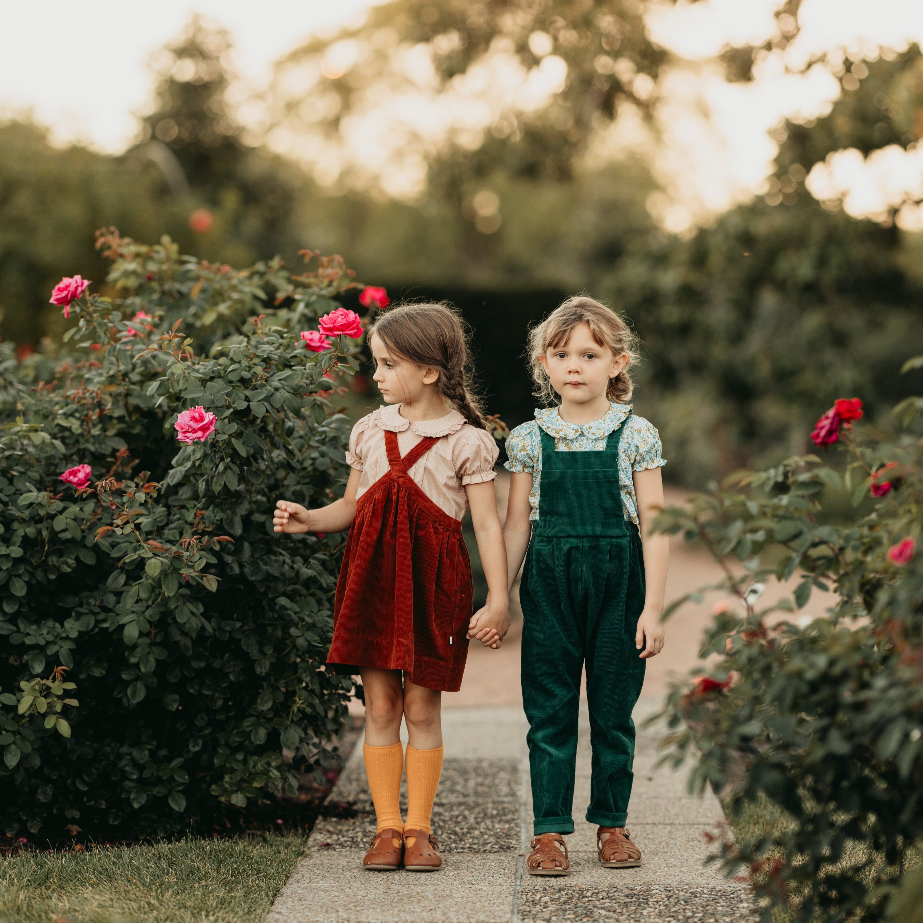 Two girls holding hands in a garden with pink flowers and greenery.