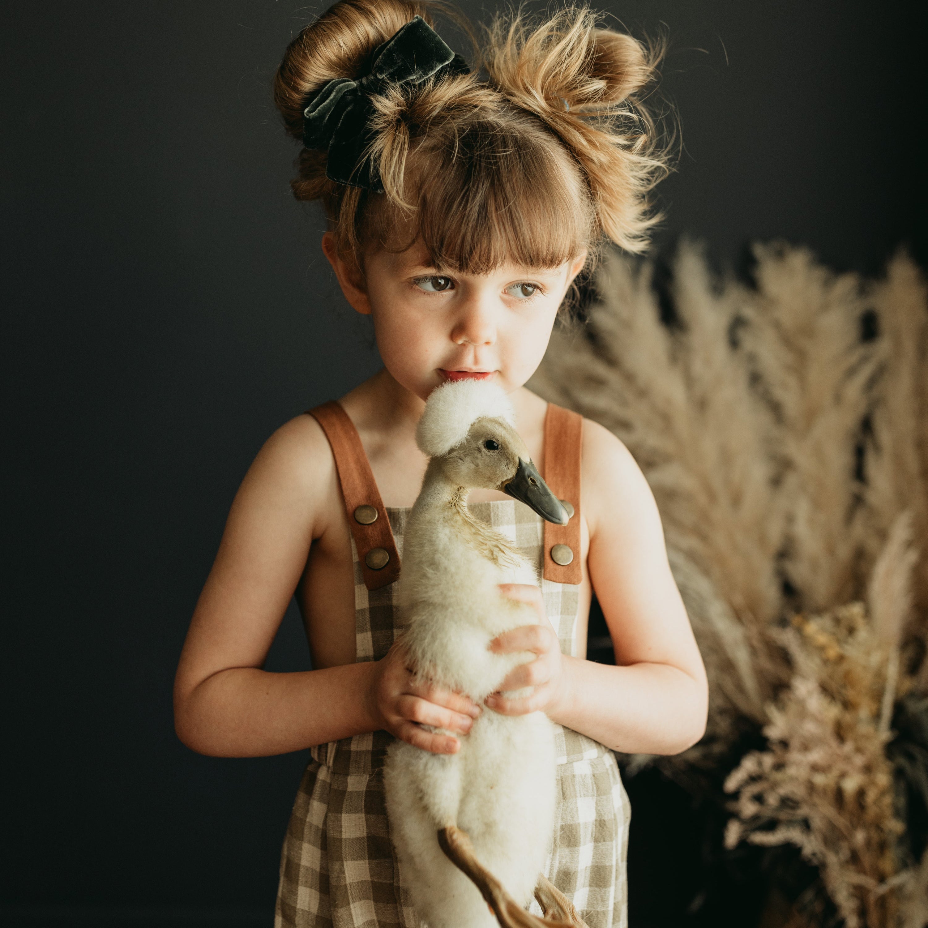 Little 5-year-old girl holding a goose with love, wearing beige gingham linen long overalls featuring a gender-neutral design, breathable fabric for comfort, stylish pleats, durable leather-looking straps, adjustable straps for a perfect fit, and unfoldable leg cuffs designed to grow with her.