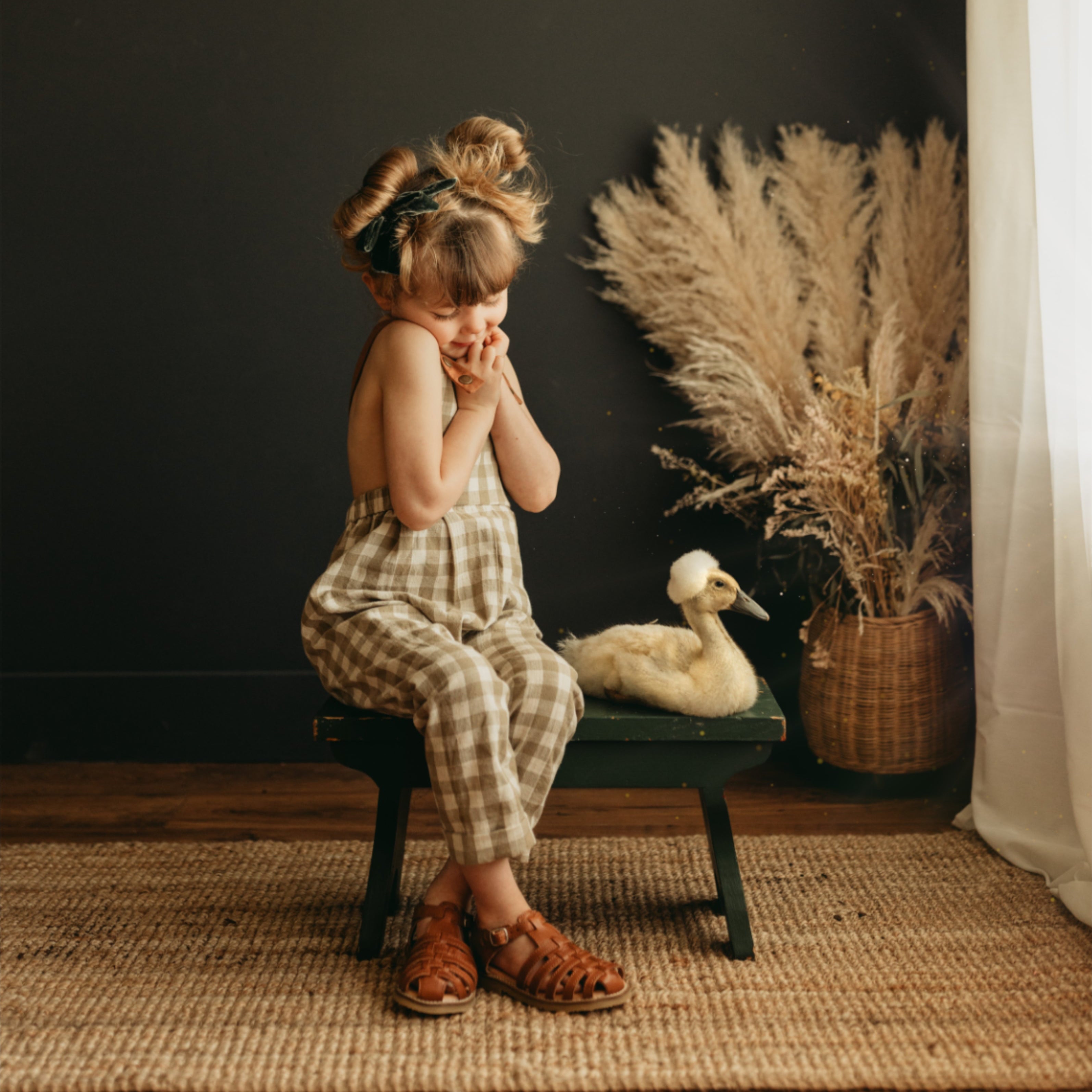 Little 5-year-old girl sitting indoors next to a goose, wearing beige gingham linen long overalls featuring a gender-neutral design, breathable fabric for comfort, stylish pleats, durable leather-looking straps, adjustable straps for a perfect fit, and unfoldable leg cuffs designed to grow with her.