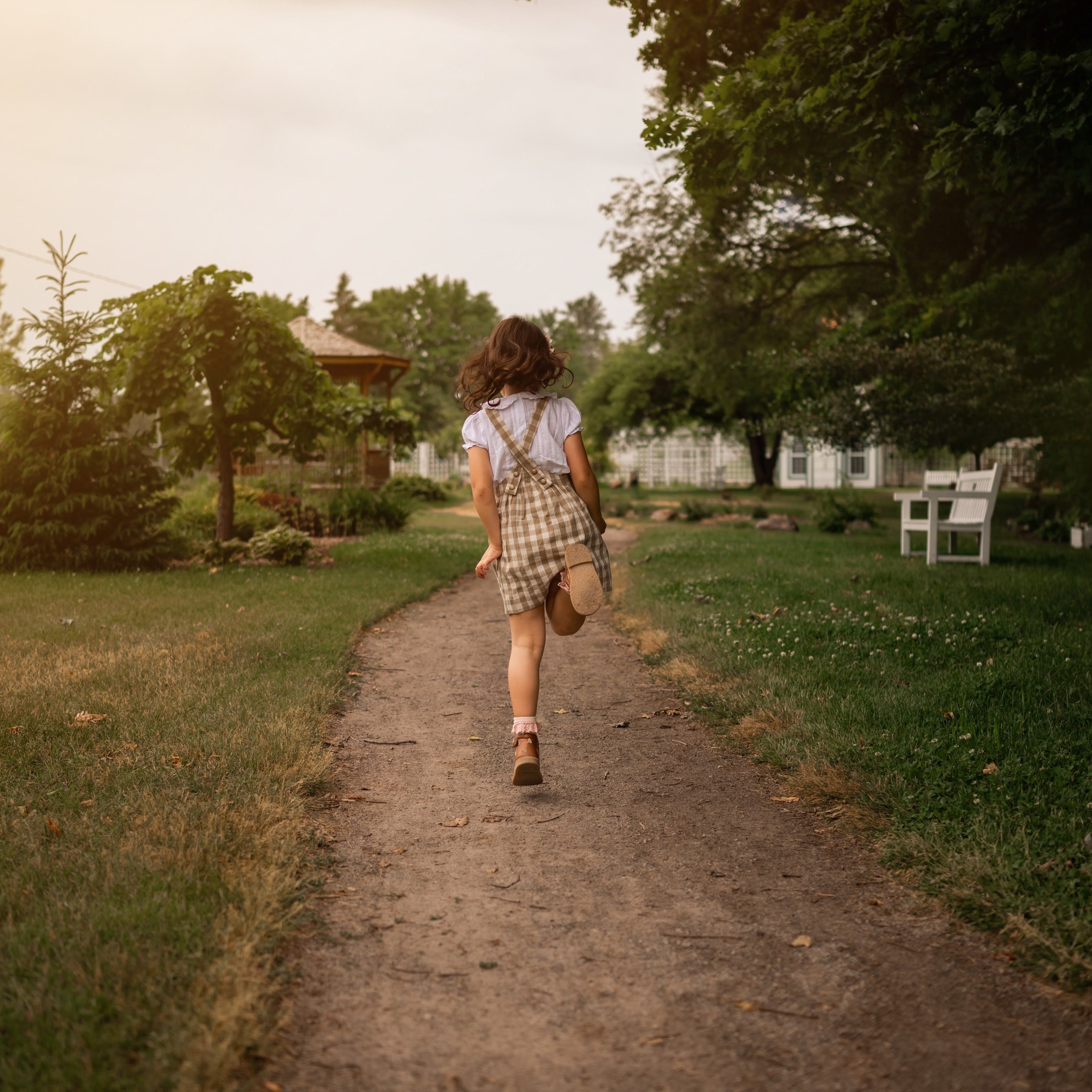 Petite 6-year-old girl seen from the back, skipping playfully while wearing the Beige Gingham Pinafore and a white vintage heirloom style blouse, capturing the carefree spirit and joy of childhood in her stylish outfit.