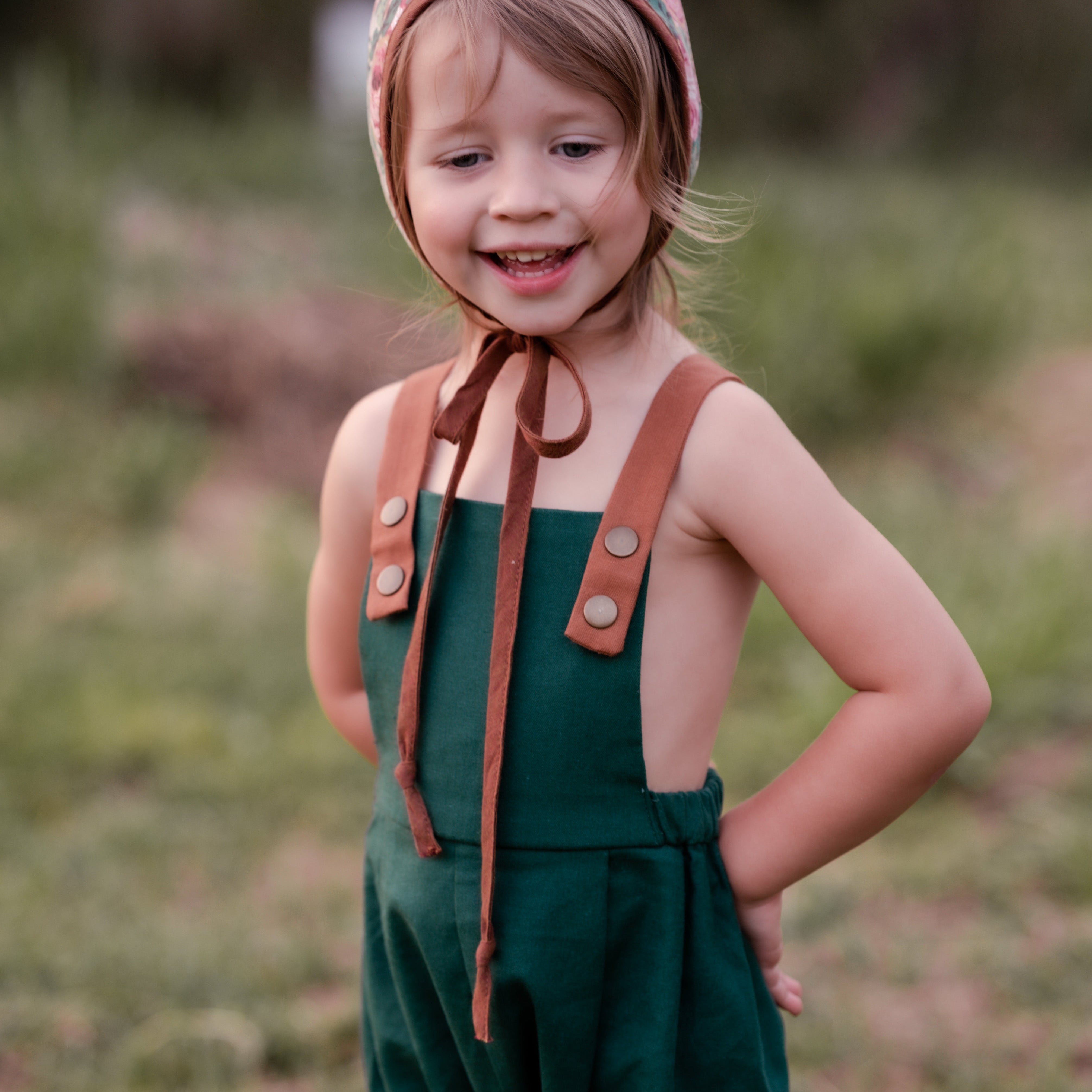 A 3-year-old girl standing and laughing in evergreen long overalls, capturing the joy and playful spirit of young children in stylish clothing.