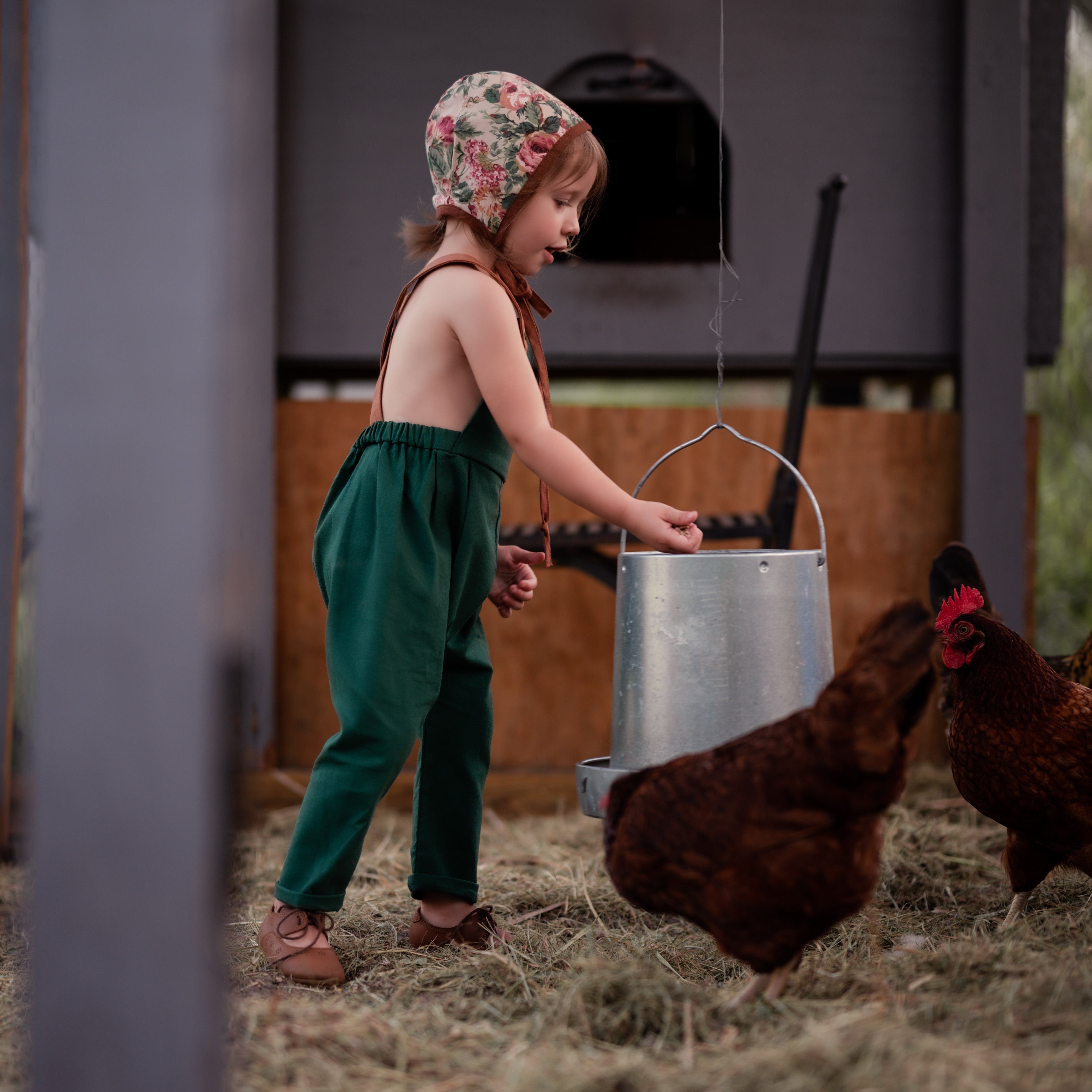 Side view of a 3-year-old girl in the barn wearing evergreen linen long overalls, showcasing the fit and elegant design details that make these overalls a favorite.