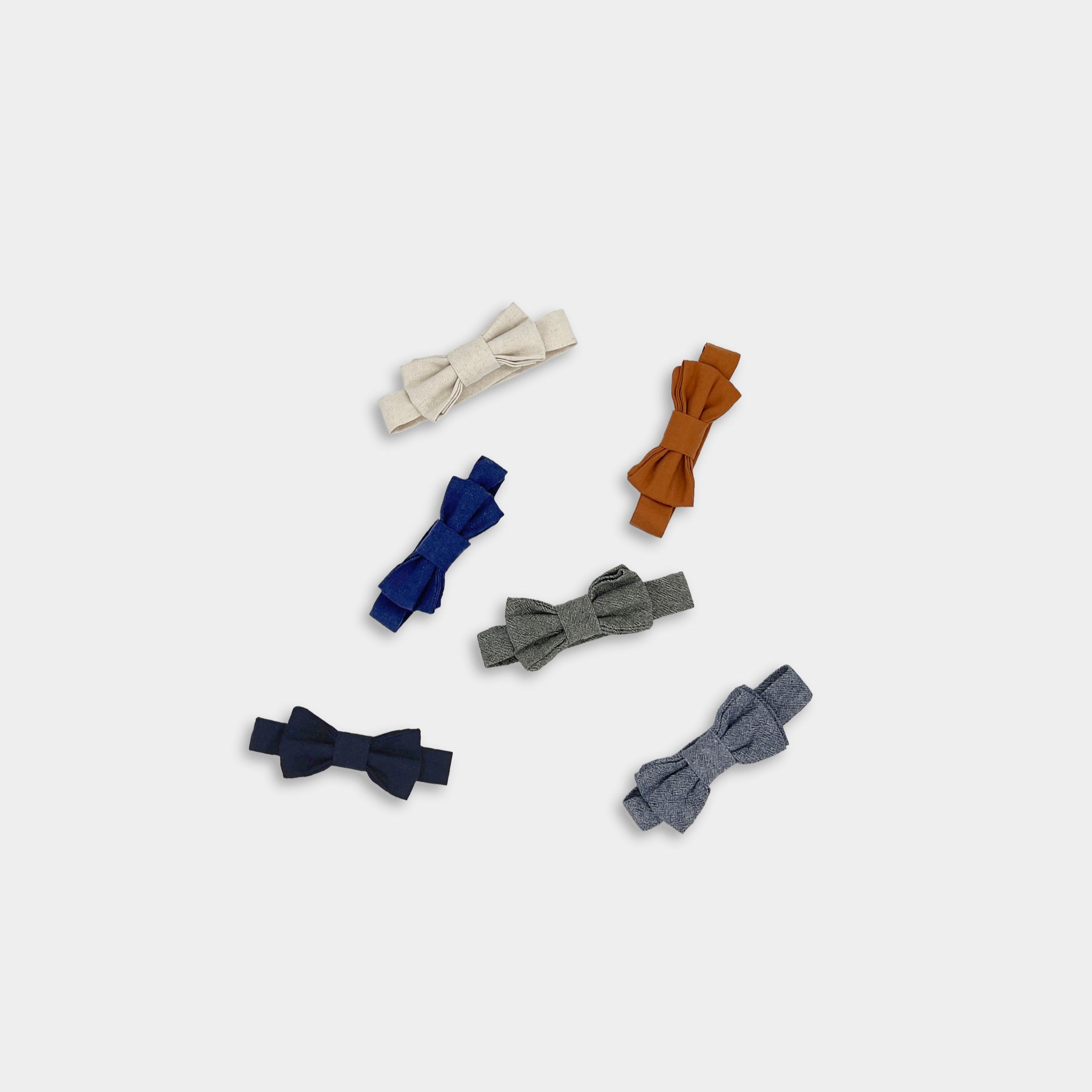 Photo showcasing all six linen bow ties in a row: Navy, Black, Natural, Burnt Orange, Chambray Blue, and Olive Green - This vibrant display highlights the variety of colors available, emphasizing the timeless design and versatility of each bow tie. The arrangement allows for a clear comparison of the colors, making it easy to choose the perfect accessory to complement any outfit.