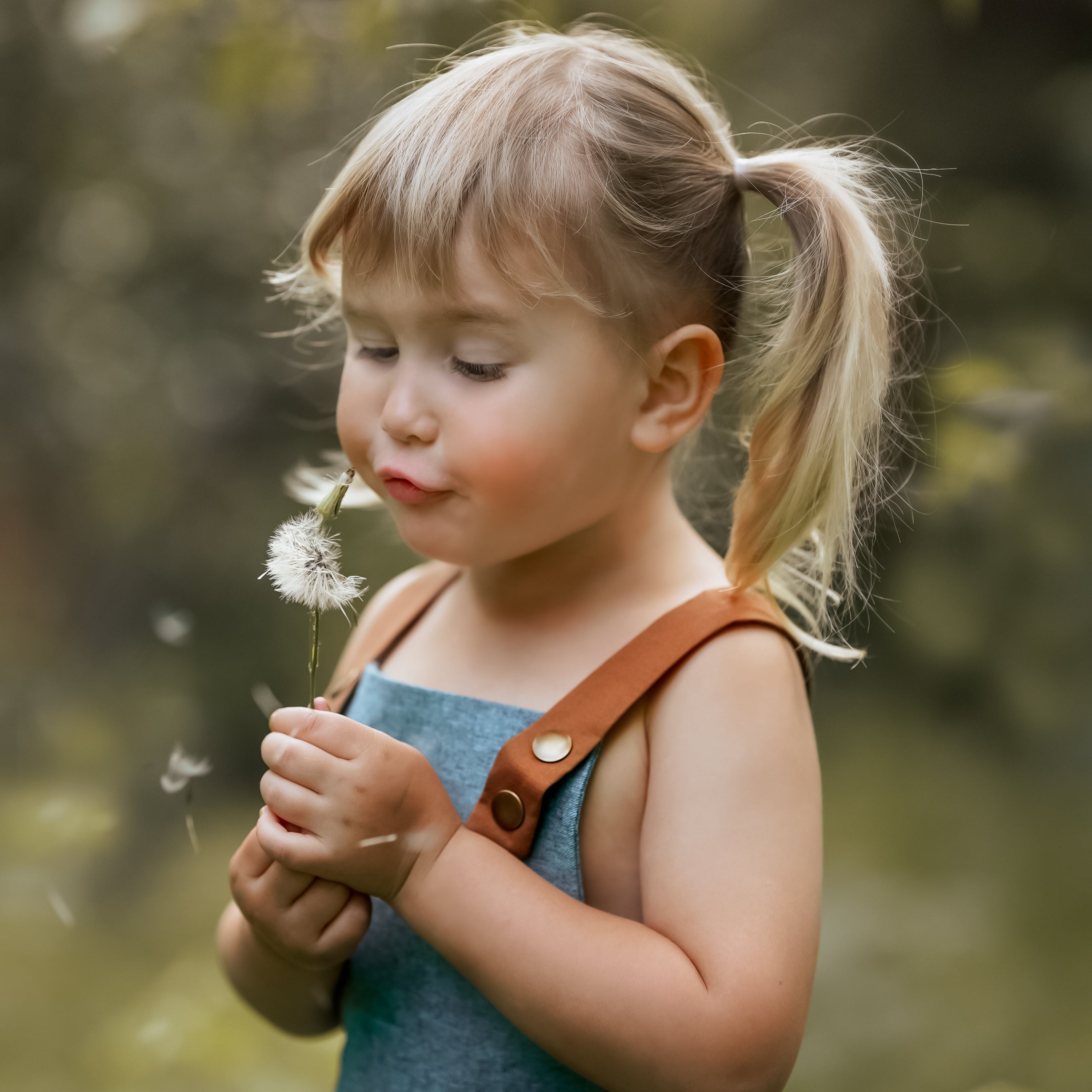 A close-up of a little girl in a Blue Jay bubble romper, joyfully blowing dandelion seeds into the air. Her face is filled with delight, capturing the essence of childhood wonder and the playful spirit of summer.
