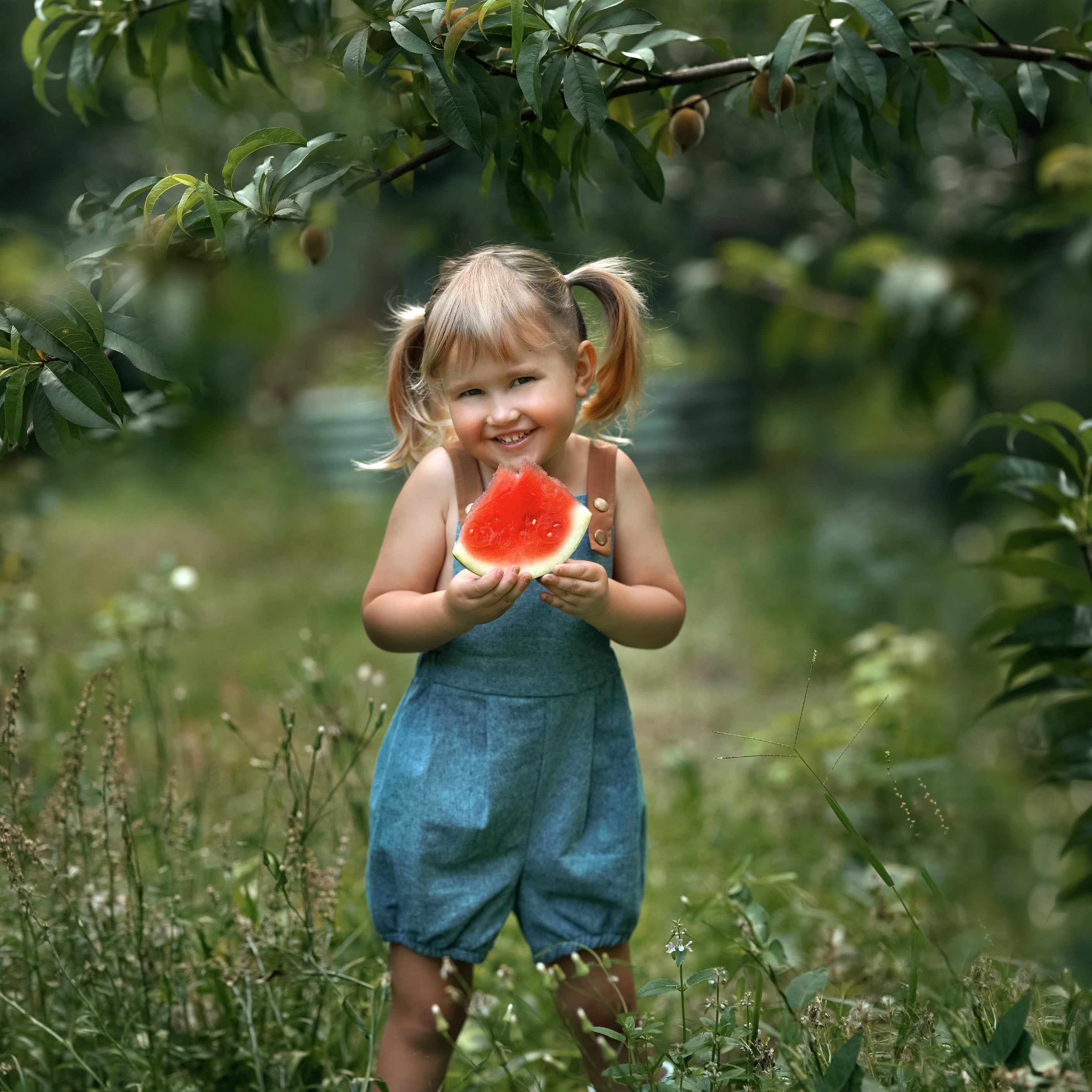 A little girl wearing a Blue Jay bubble romper, smiling brightly while enjoying a slice of watermelon. The romper's adjustable straps and elasticized legs complement her playful demeanor, making it perfect for a summer day.