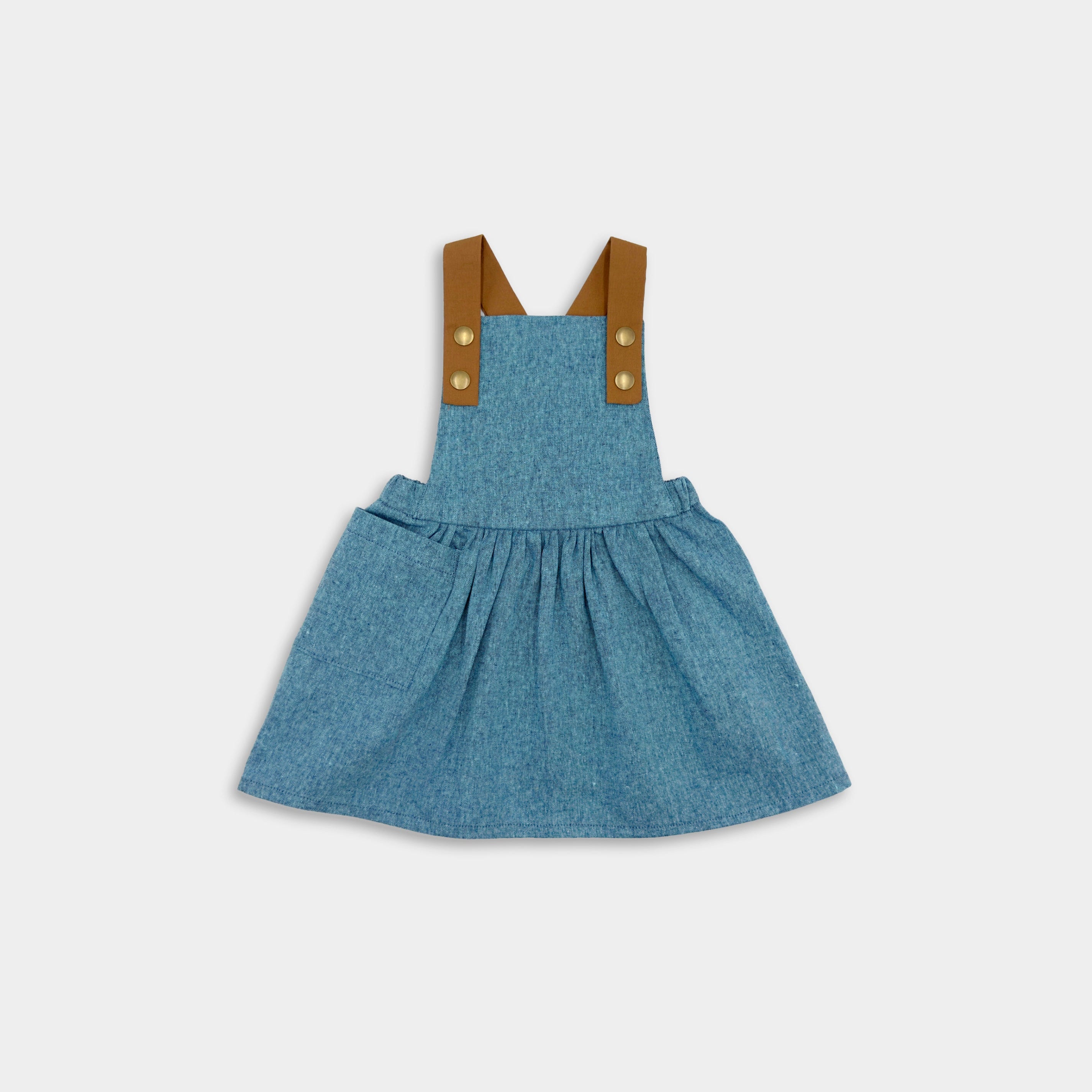 Pinafore in Blue Jay on a little girl model enjoying summer - This classic and timeless design is handmade in our NYC studio using repurposed linen. The pinafore is designed to be worn on its own, ensuring your little girl stays stylish and cool all summer long. Durable leather-looking straps enhance visual appeal, while adjustable straps in two lengths provide a perfect fit. The generous pocket is perfect for storing her little treasures during outdoor adventures.