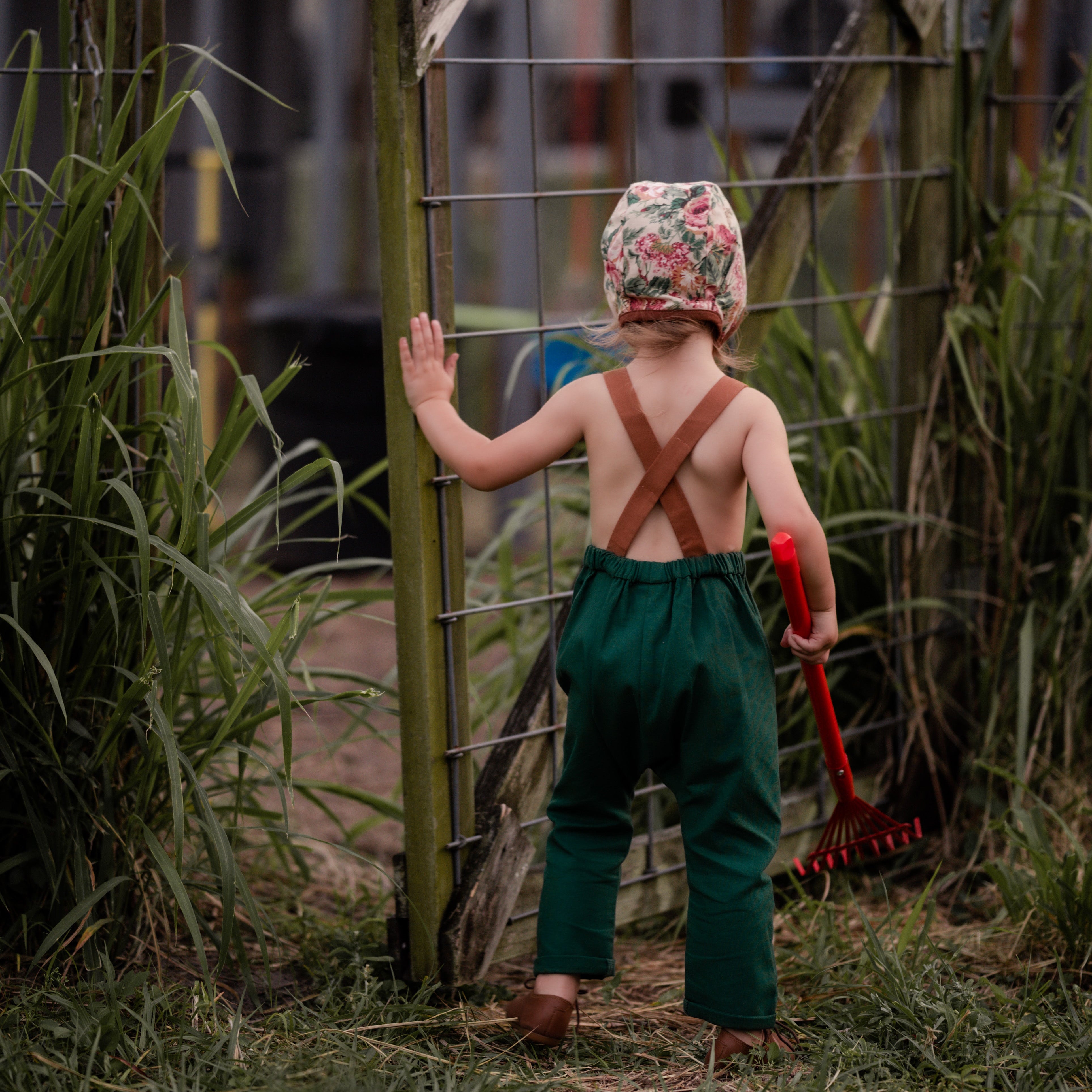Back view of a 3-year-old girl closing a wooden gate in evergreen linen long overalls, highlighting the adjustable straps and overall functionality for everyday wear.