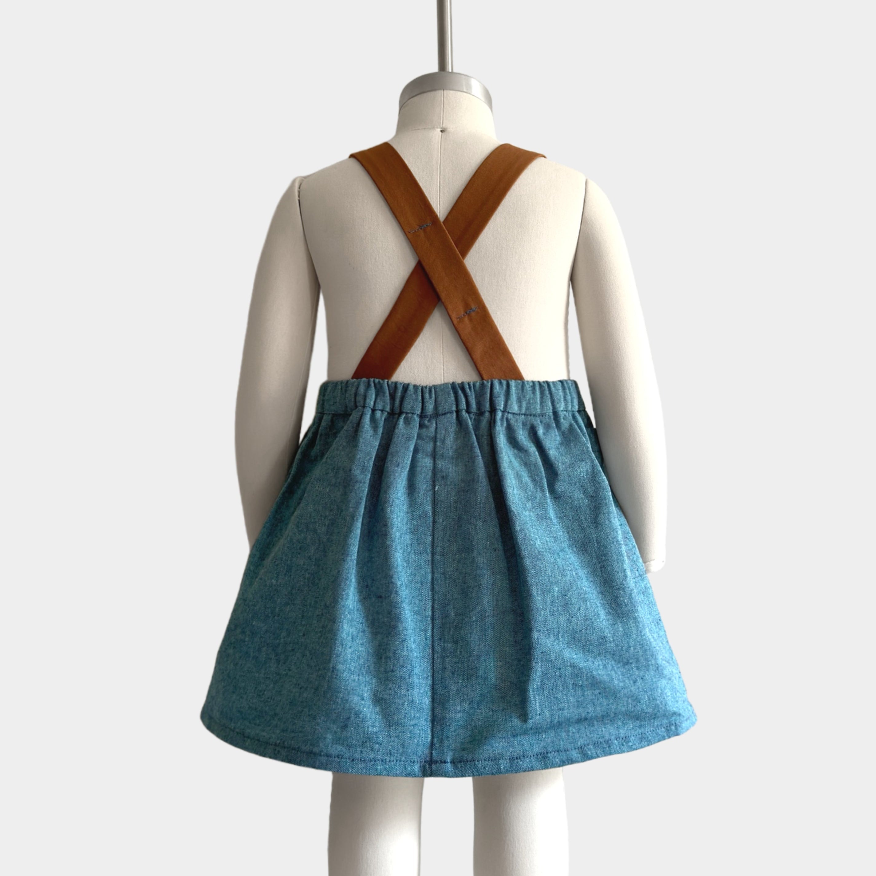 The Blue Jay pinafore displayed on a mannequin, highlighting the back view with an emphasis on the original brown leather-looking straps - This classic design, handmade from repurposed linen in our NYC studio, showcases the durability and visual appeal of the straps. The adjustable lengths ensure a perfect fit, while the generous pocket adds practicality, making this piece a stylish choice for summer wear.