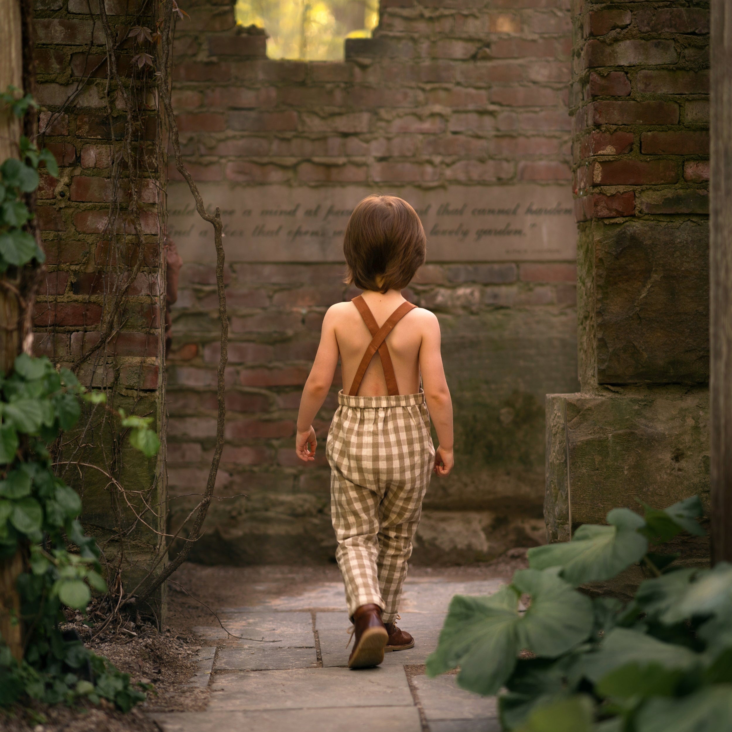 5-year-old boy wearing beige gingham linen long overalls walking away from the camera, showcasing the back view of the overalls in a playful outdoor setting. Overalls with brown contrasting straps.
