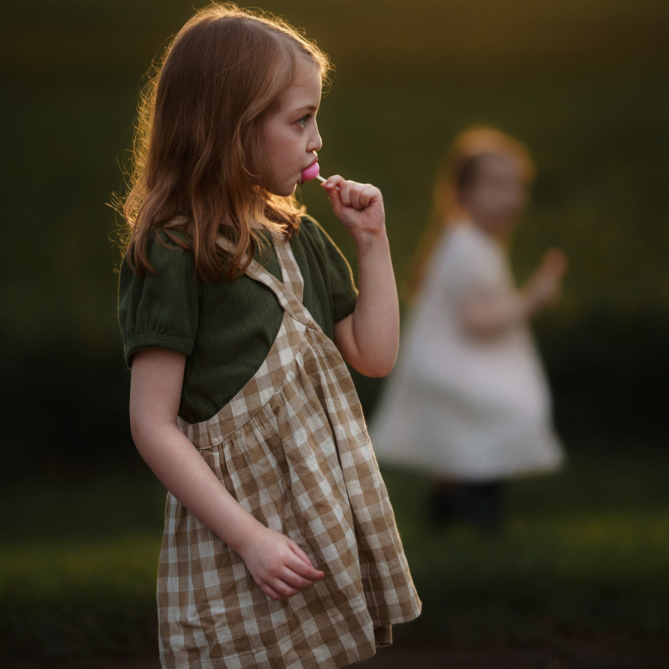 Young girl in a checkered dress standing in a field with a blurred figure in the background during sunset.