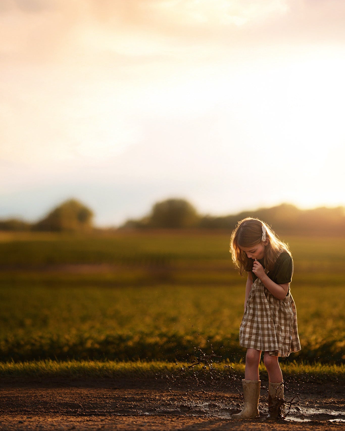 Young girl standing in a field at sunset, splashing in a puddle with rainbows eating a lollipop dressed in a checkered pinafore