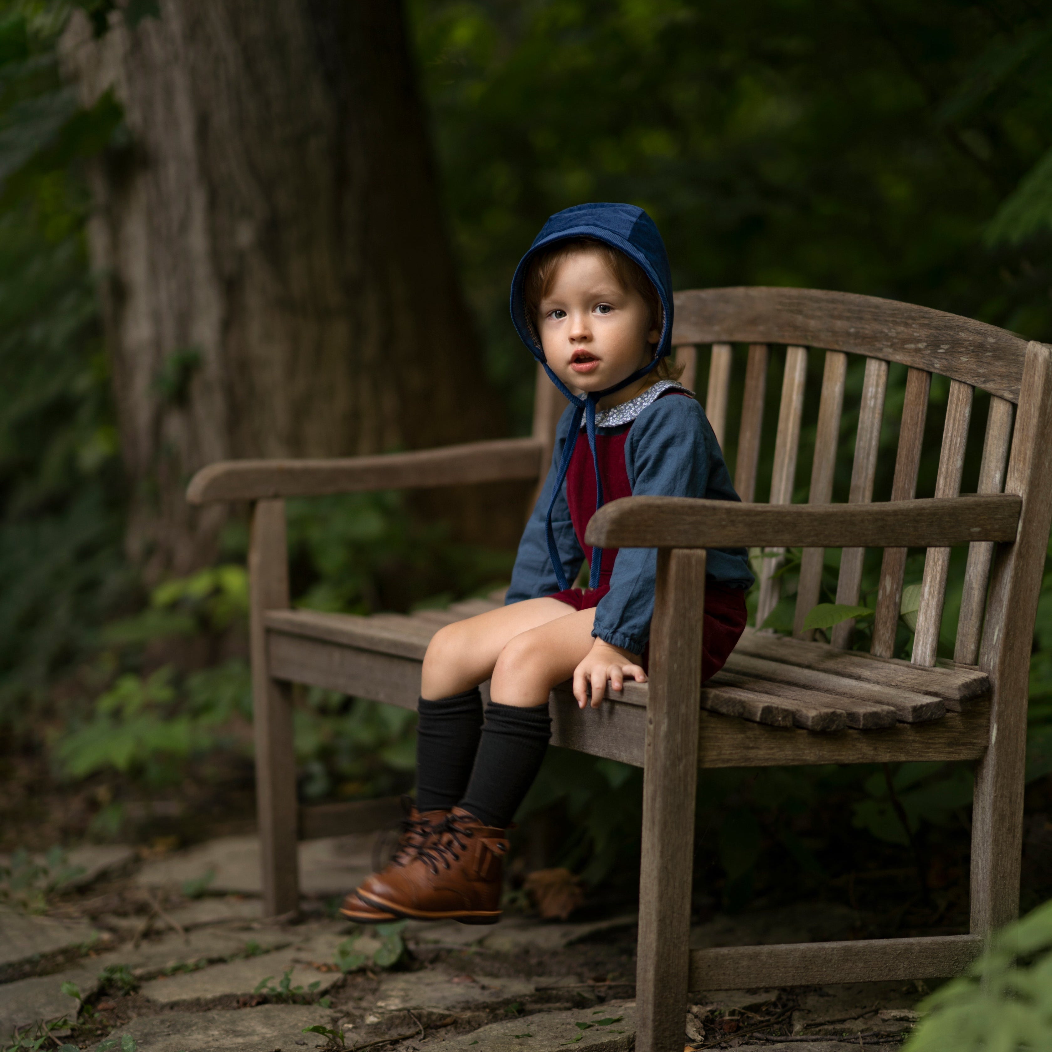 A 4-year-old boy sitting on a bench while wearing the cornflower indigo blue corduroy bonnet, highlighting its timeless accessory appeal and comfort for everyday wear.