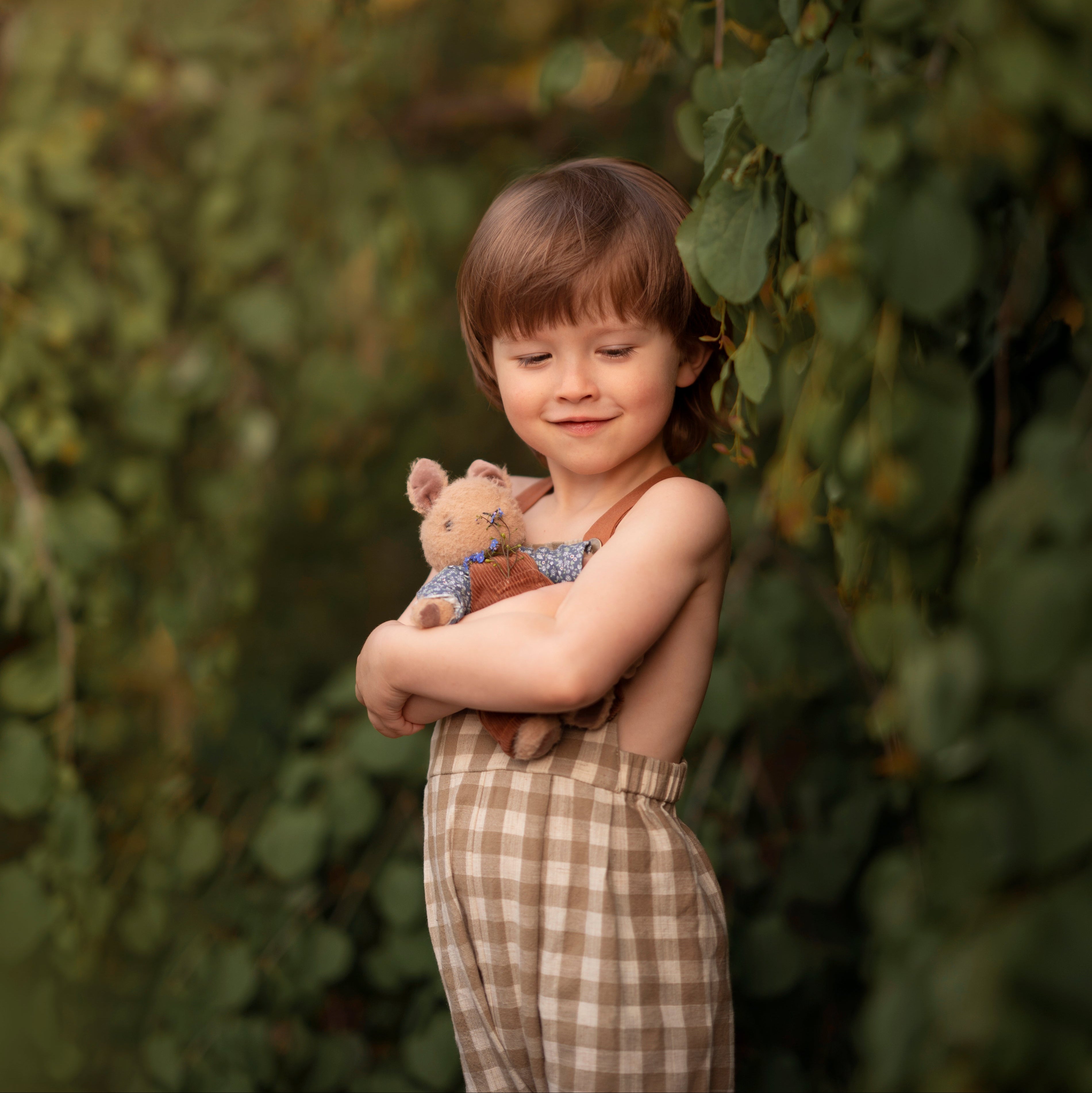5-year-old boy wearing beige gingham linen long overalls while holding a teddy bear, featuring a gender-neutral design, breathable fabric for comfort, and adjustable straps for a perfect fit.