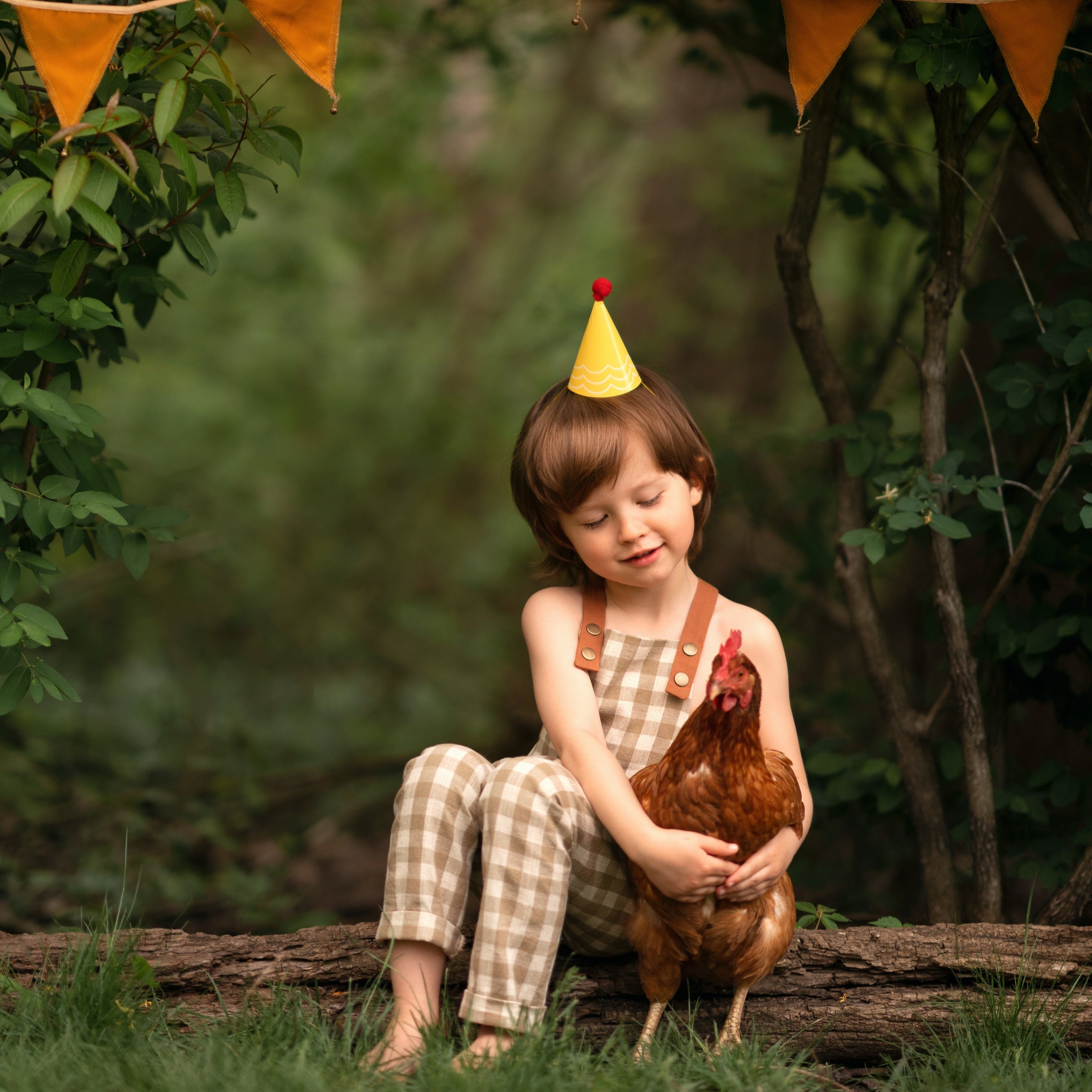 5-year-old boy wearing beige gingham linen long overalls in a birthday scene outdoors, surrounded by festive decorations and a hen, showcasing a gender-neutral design and breathable fabric for comfort.