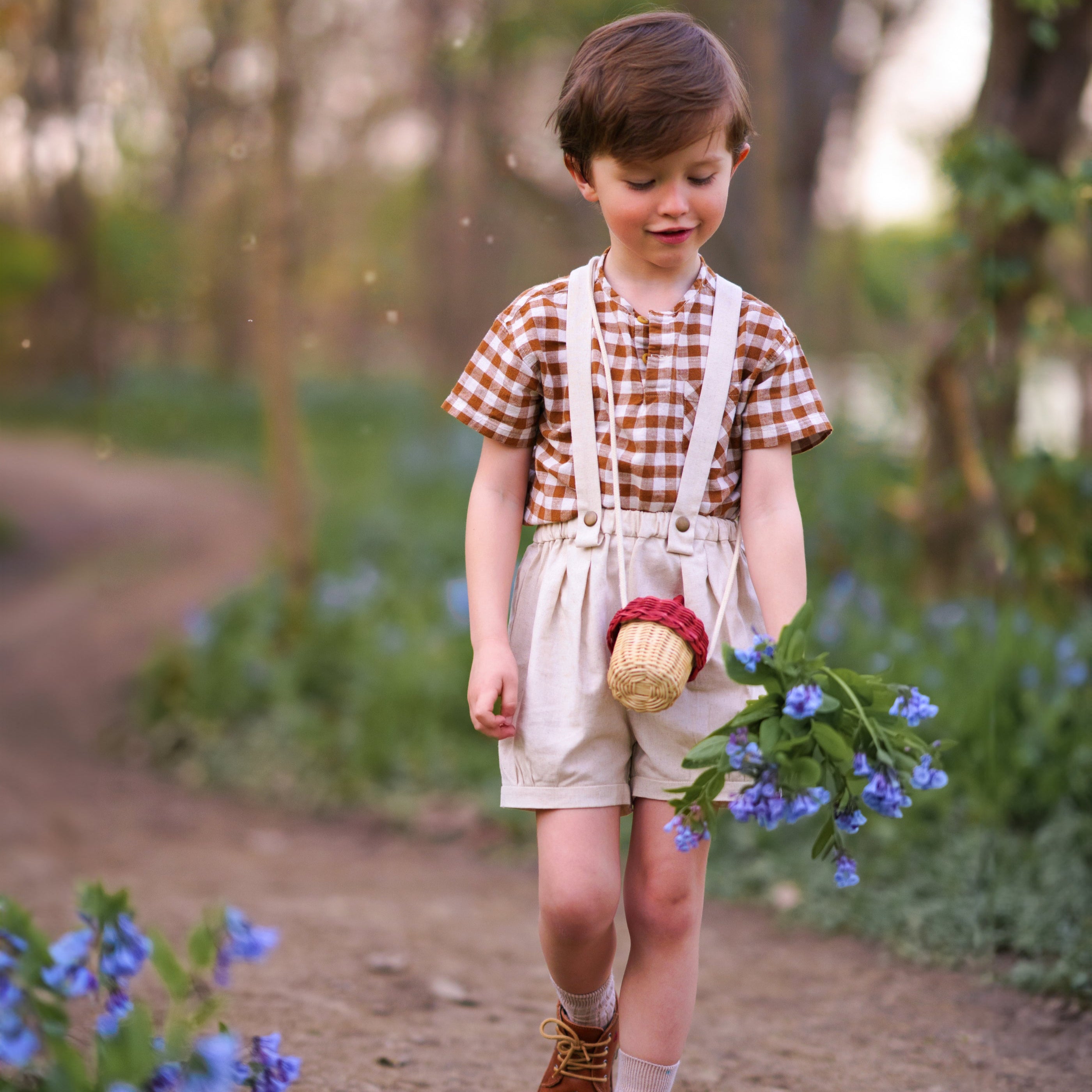 Child in a checkered shirt and overalls holding flowers in a forest setting