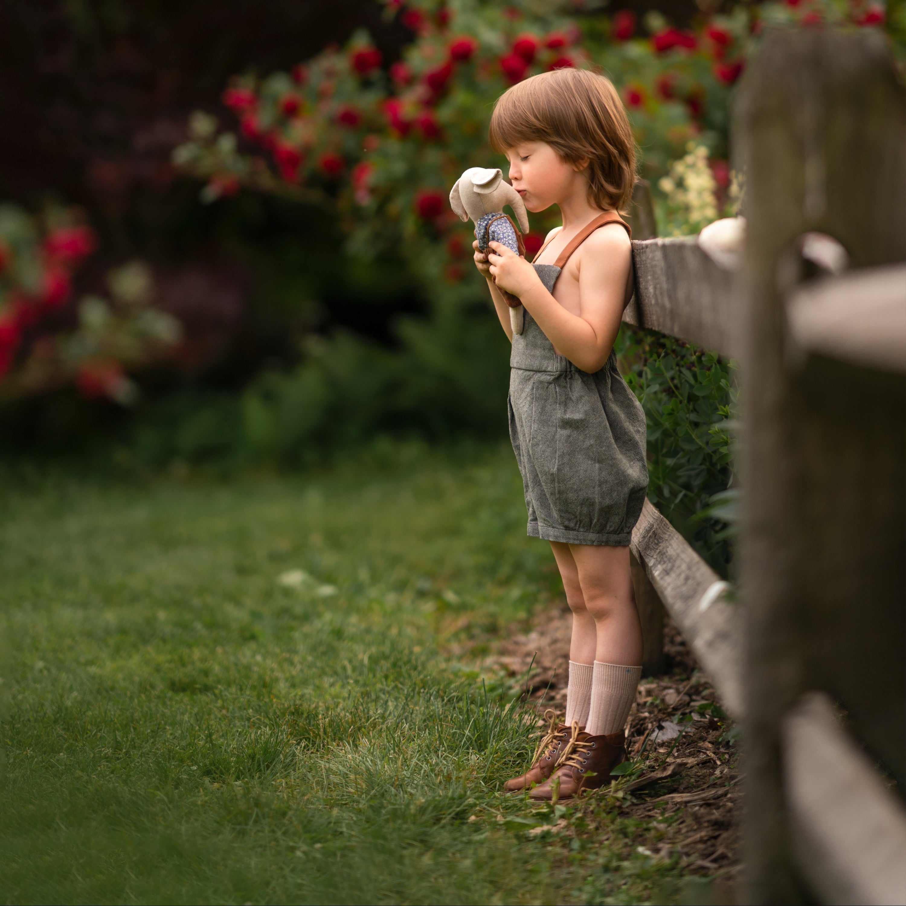 Young boy in green overalls holding a teddy bear in a garden setting